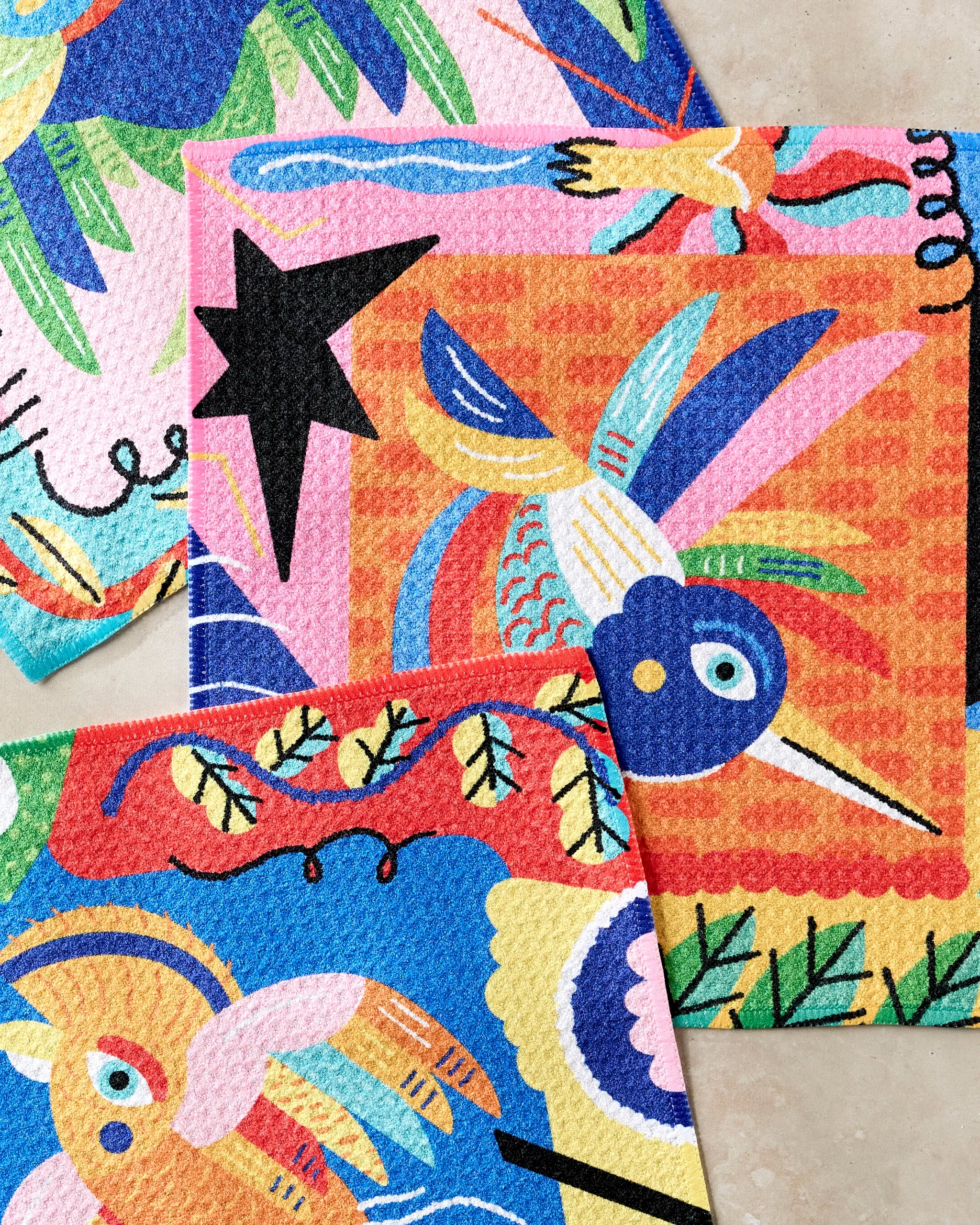 Camila Leão’s Tropical Birds set includes three eco-friendly cloths in bold blue, pink, orange, yellow, green, and black with vibrant abstract bird and plant designs on a light beige background.