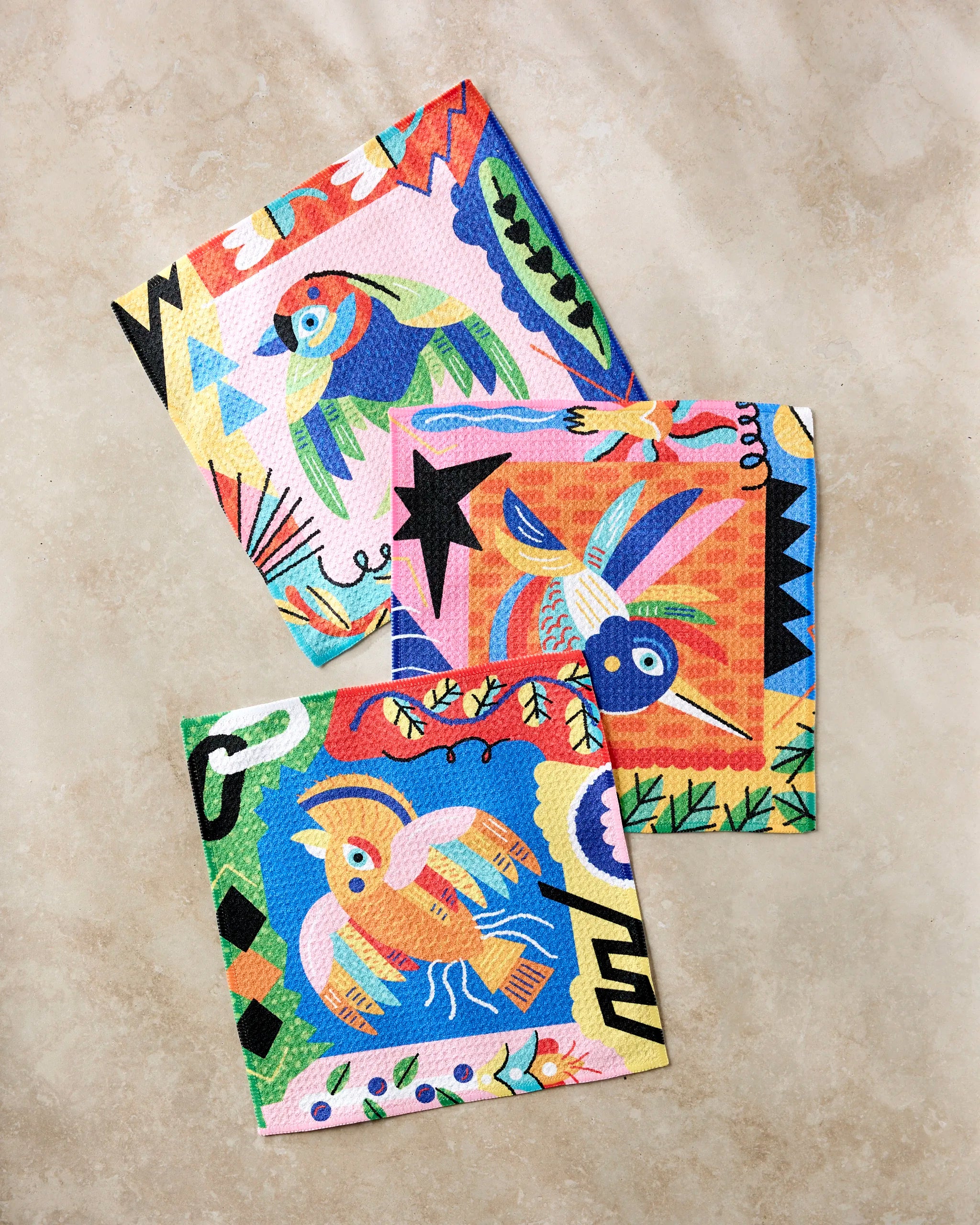 Three Tropical Birds napkins by Camila Leão, each with a unique abstract bird illustration and decorative borders, are crafted from recycled materials and displayed on a beige marble surface.