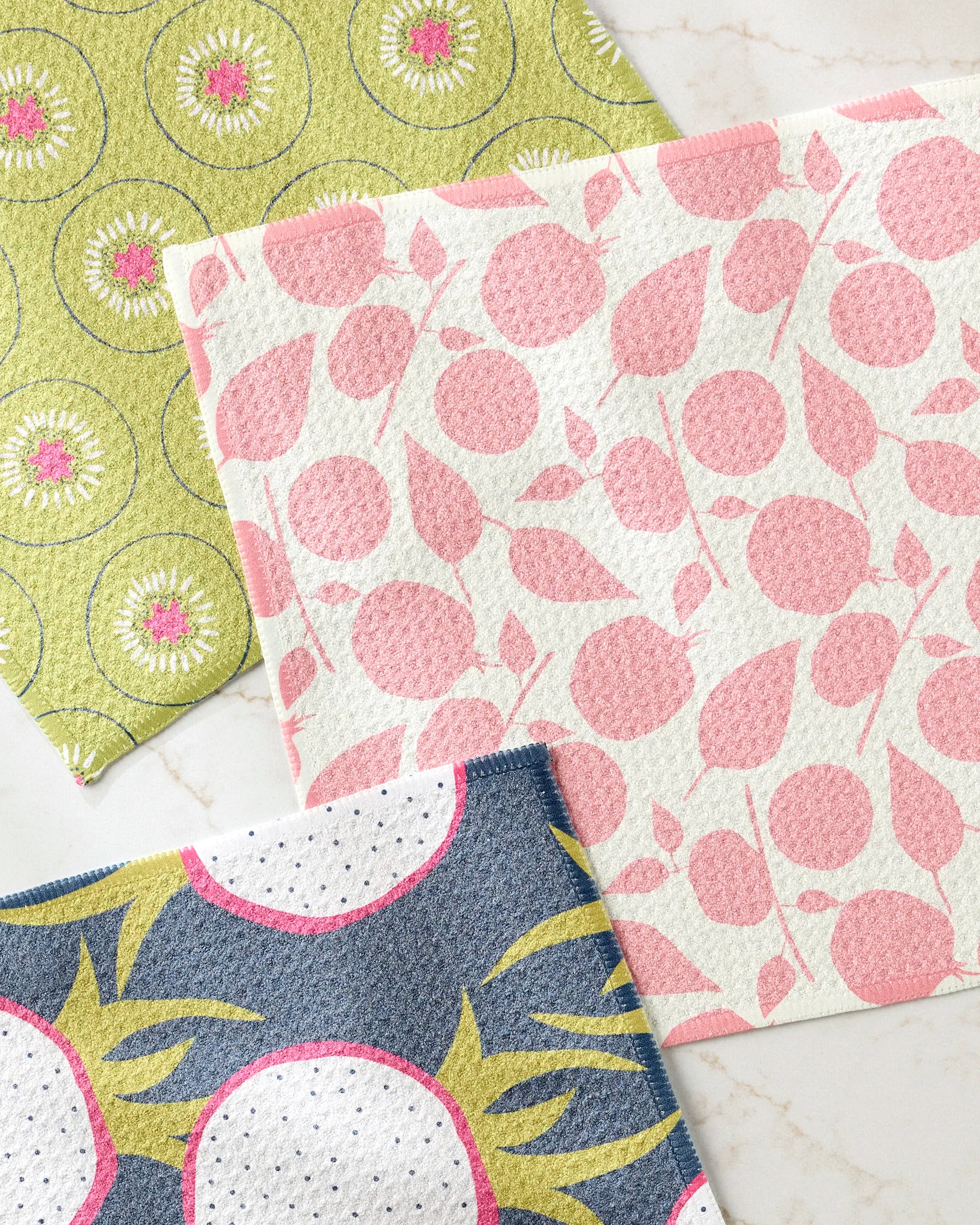 Three cloths from the Lizzie Clark "Tropical Fruit" set are arranged on a marble surface: one with pink leaves and circles, one green with floral designs, and one blue with white circles and yellow sunbursts.