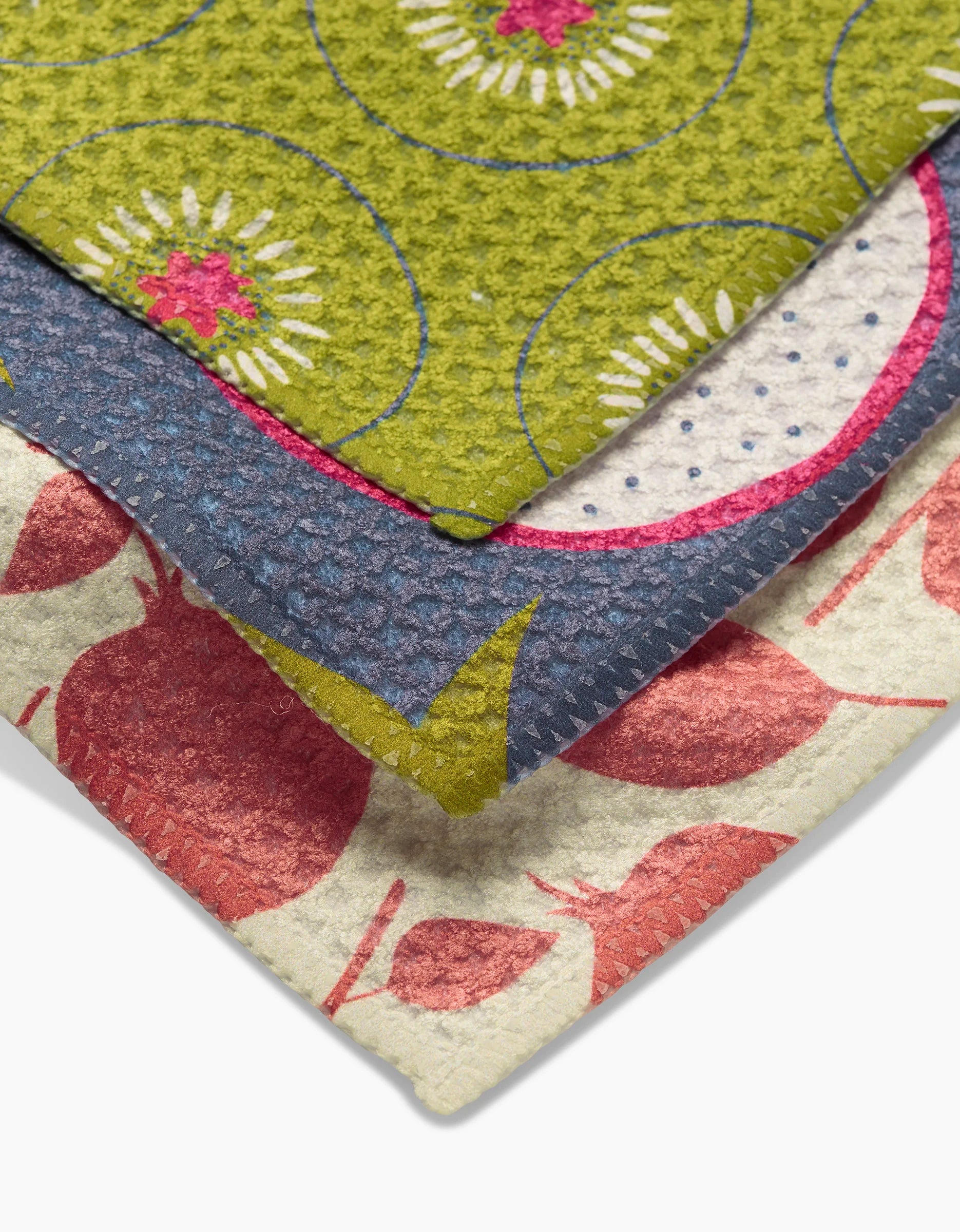 Three textured fabric pieces from the Lizzie Clark Tropical Fruit Dishtowel Set feature colorful patterns: green with circular floral designs, blue with polka dots and pink accents, and cream with red fruit-like motifs.