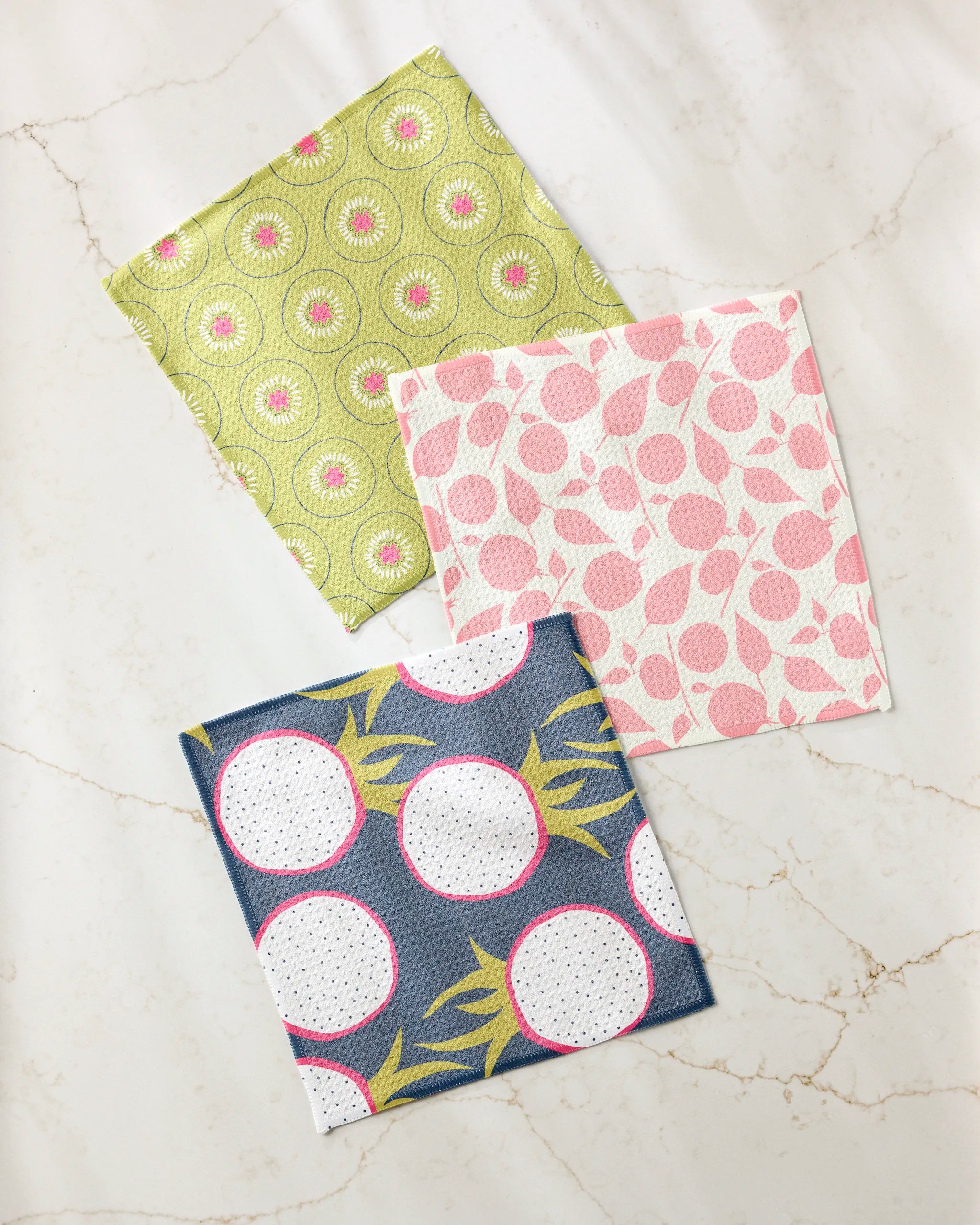 The Lizzie Clark Tropical Fruit dishcloth set includes three patterned cloths: green with circles, pink with abstract shapes, and dark blue with large white circles and yellow accents, displayed on a light marbled surface.