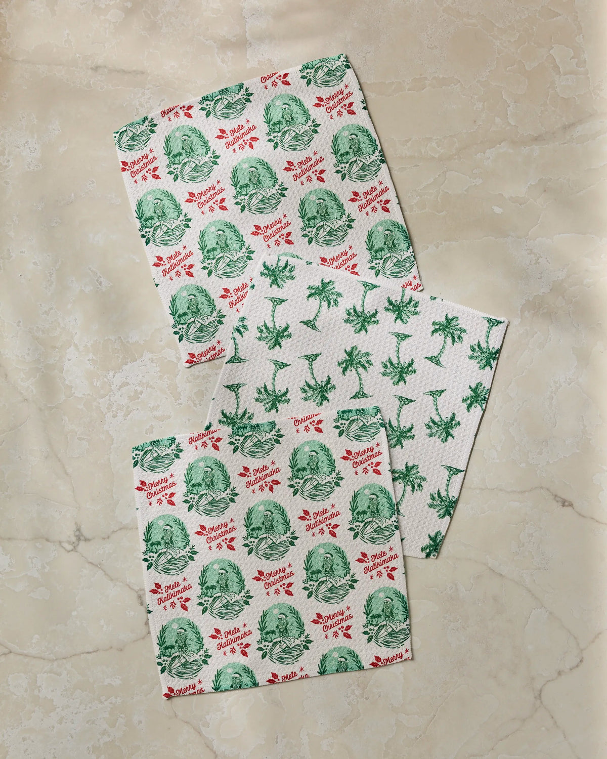 Three dishcloths from the Geometry Tropical Tidings set rest on a beige marble surface; two showcase festive green and red patterns, while one features a cheerful green palm tree design. Each kitchen towel is crafted from recycled materials, blending eco-friendly practicality with style.