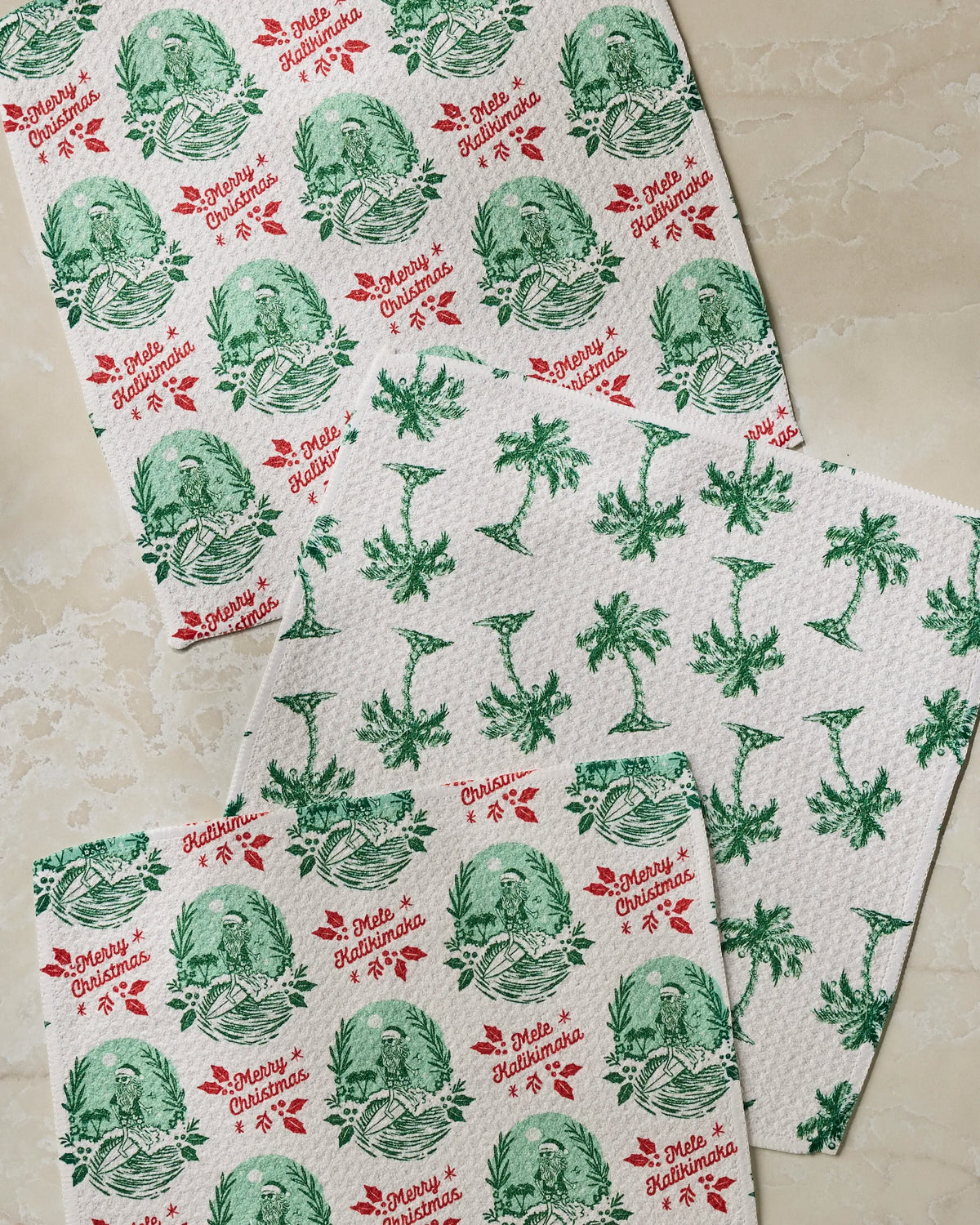 The Geometry Tropical Tidings dishcloth set includes four kitchen towels made from recycled materials, each featuring festive green and red Hawaiian-inspired Christmas patterns—such as palm trees, flowers, and the joyful messages “Mele Kalikimaka” and “Merry Christmas”—on a beige background.