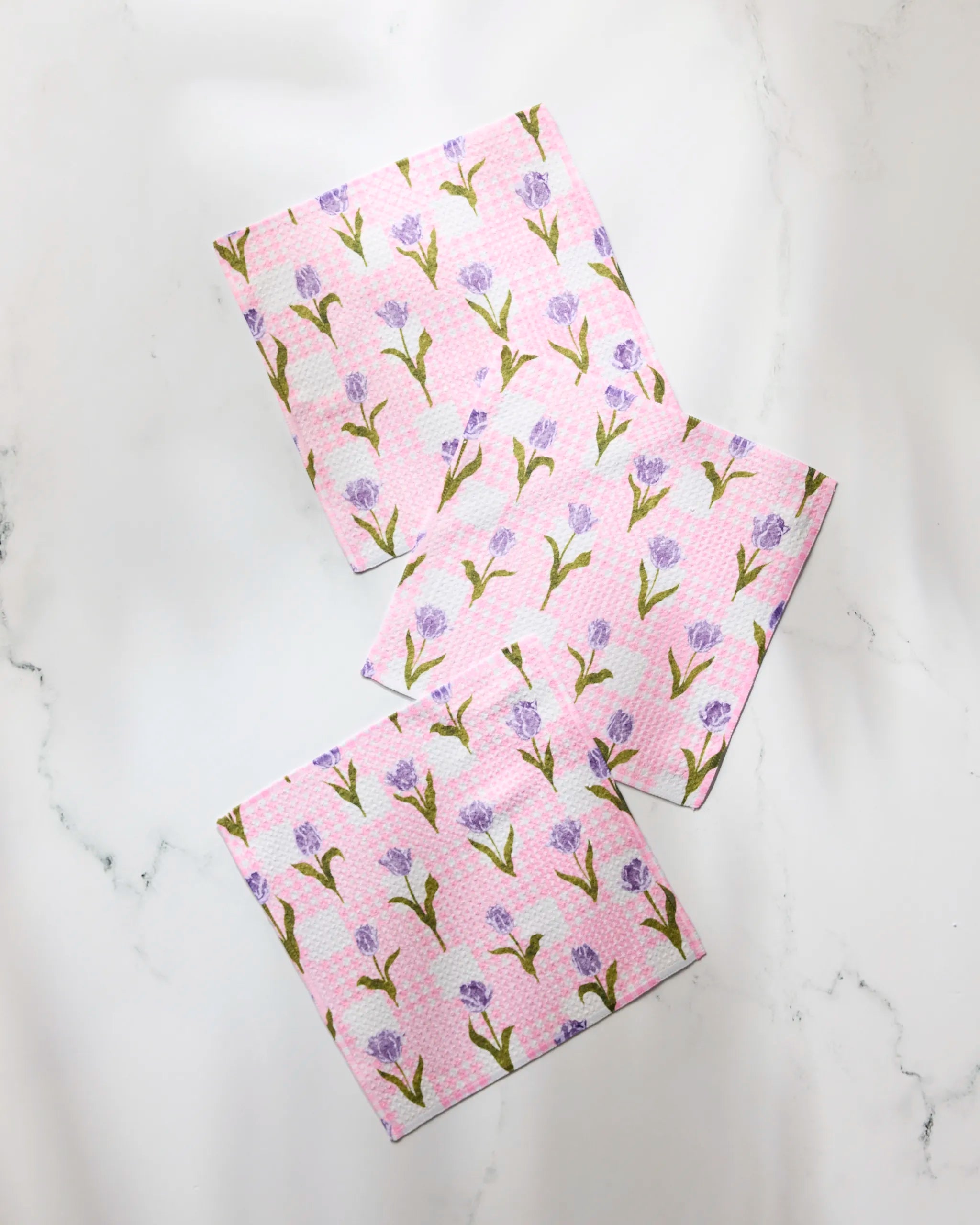 Three ‘Tulip Town’ recycled kitchen cloths by Geometry, featuring a repeating purple flower with green stems, are arranged diagonally and slightly overlapping on a white marble surface.