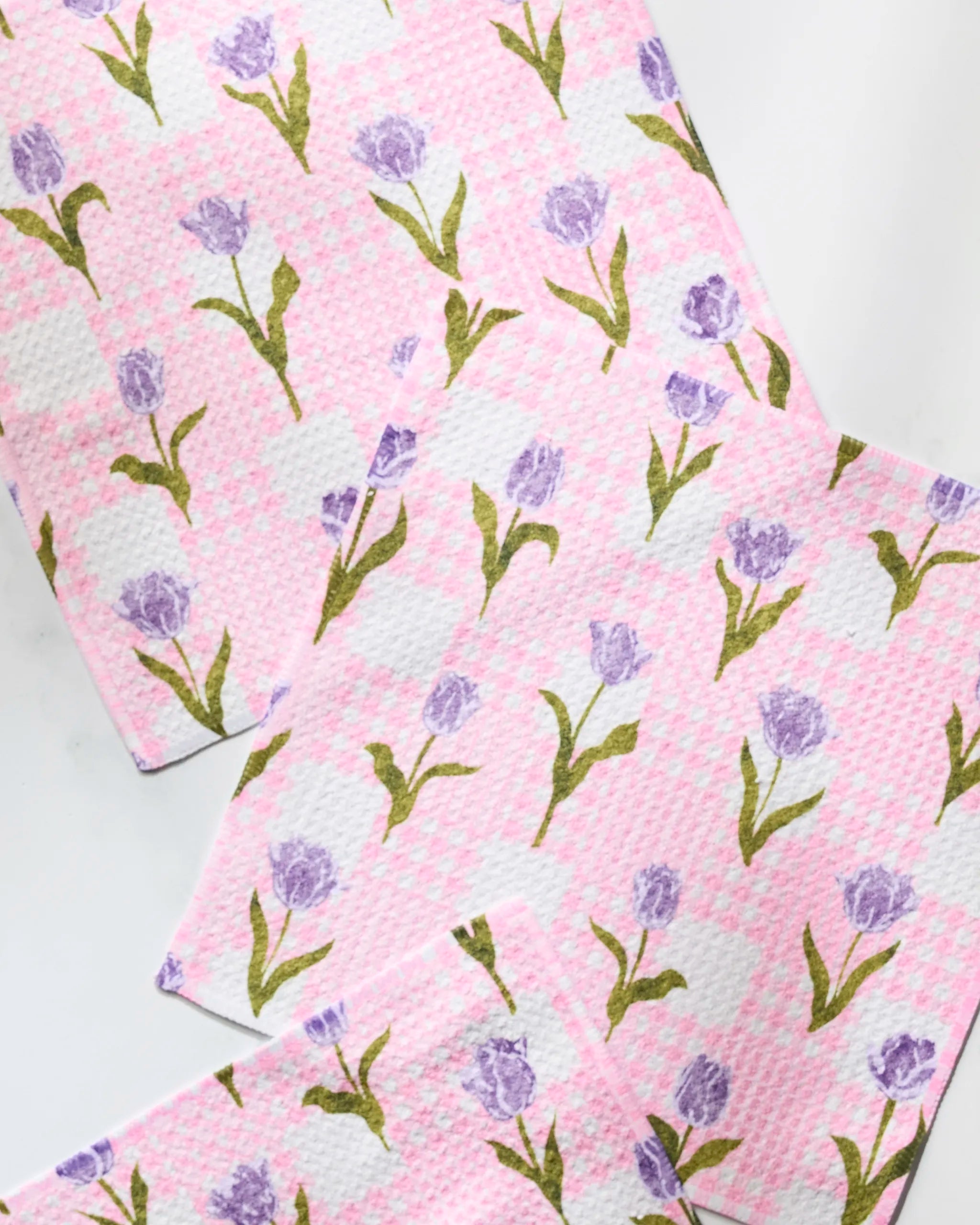 Three Tulip Town kitchen cloths by Geometry, featuring pink and white textured fabric with purple tulips and green leaves, are arranged overlapping on a white surface.