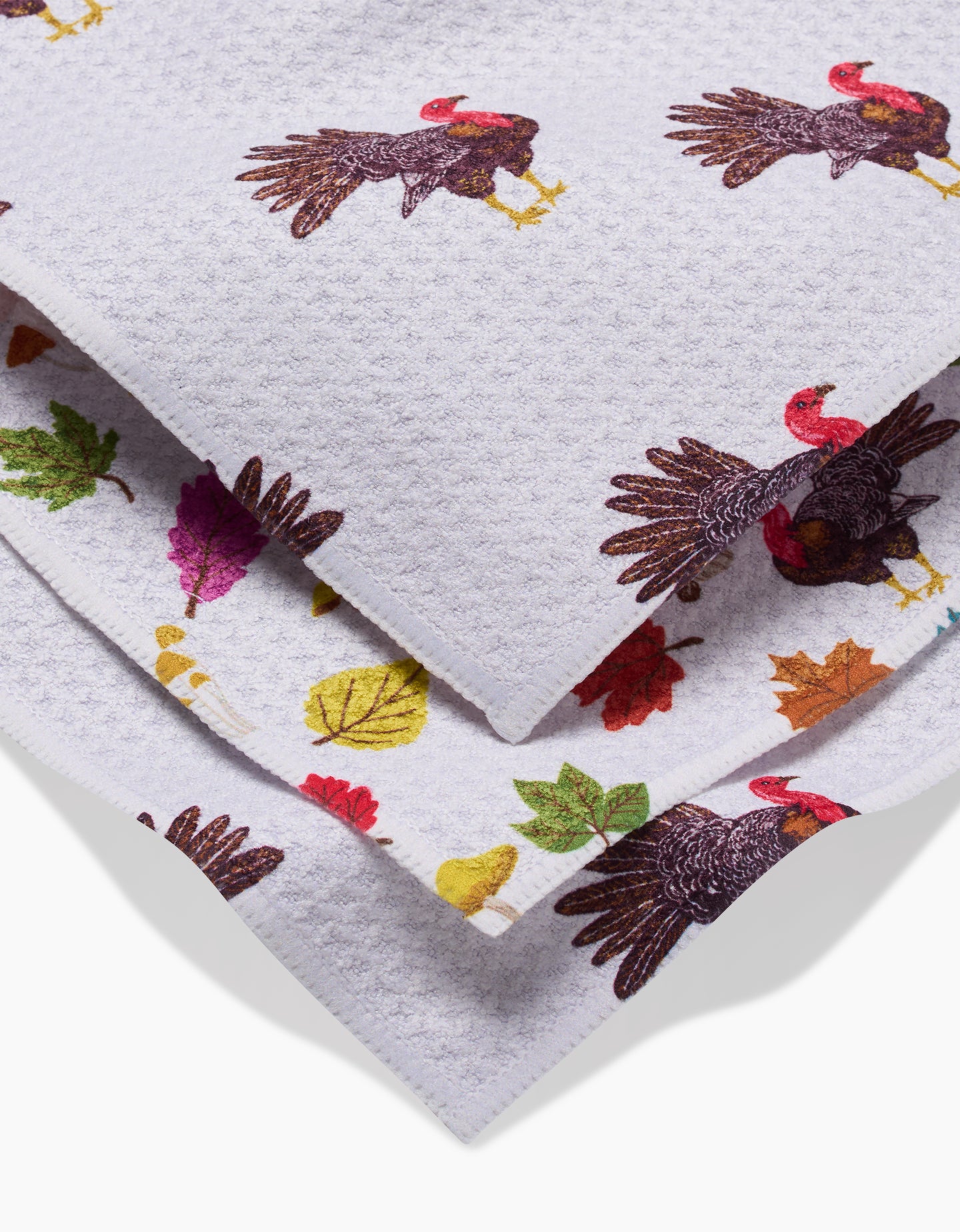 A close-up of folded white super-absorbent Turkey Day Parade dishtowels by Vaishali Design Studio, featuring colorful images of turkeys and autumn leaves printed on the fabric.