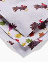 A close-up of folded white super-absorbent Turkey Day Parade dishtowels by Vaishali Design Studio, featuring colorful images of turkeys and autumn leaves printed on the fabric.