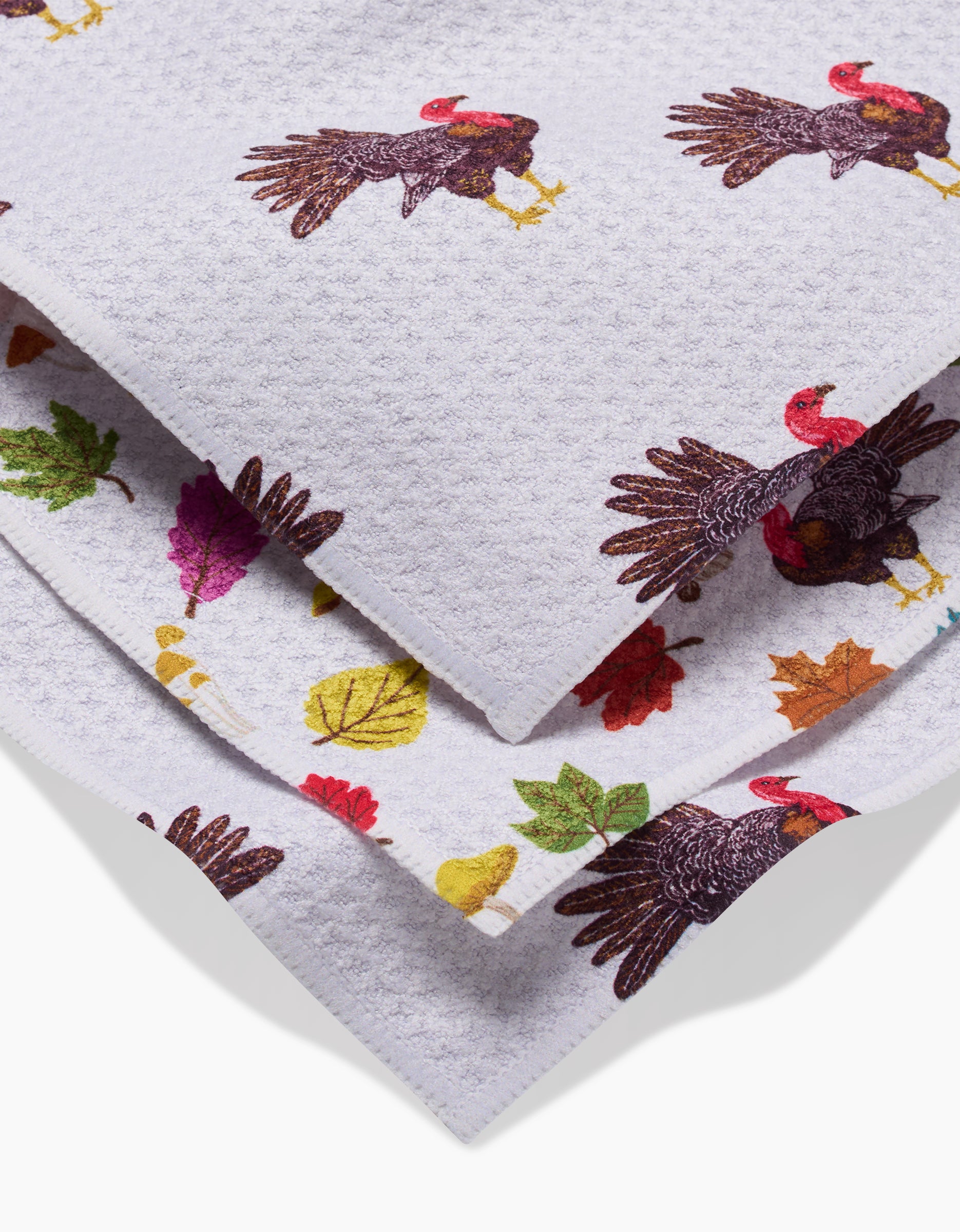 A close-up of folded white super-absorbent Turkey Day Parade dishtowels by Vaishali Design Studio, featuring colorful images of turkeys and autumn leaves printed on the fabric.
