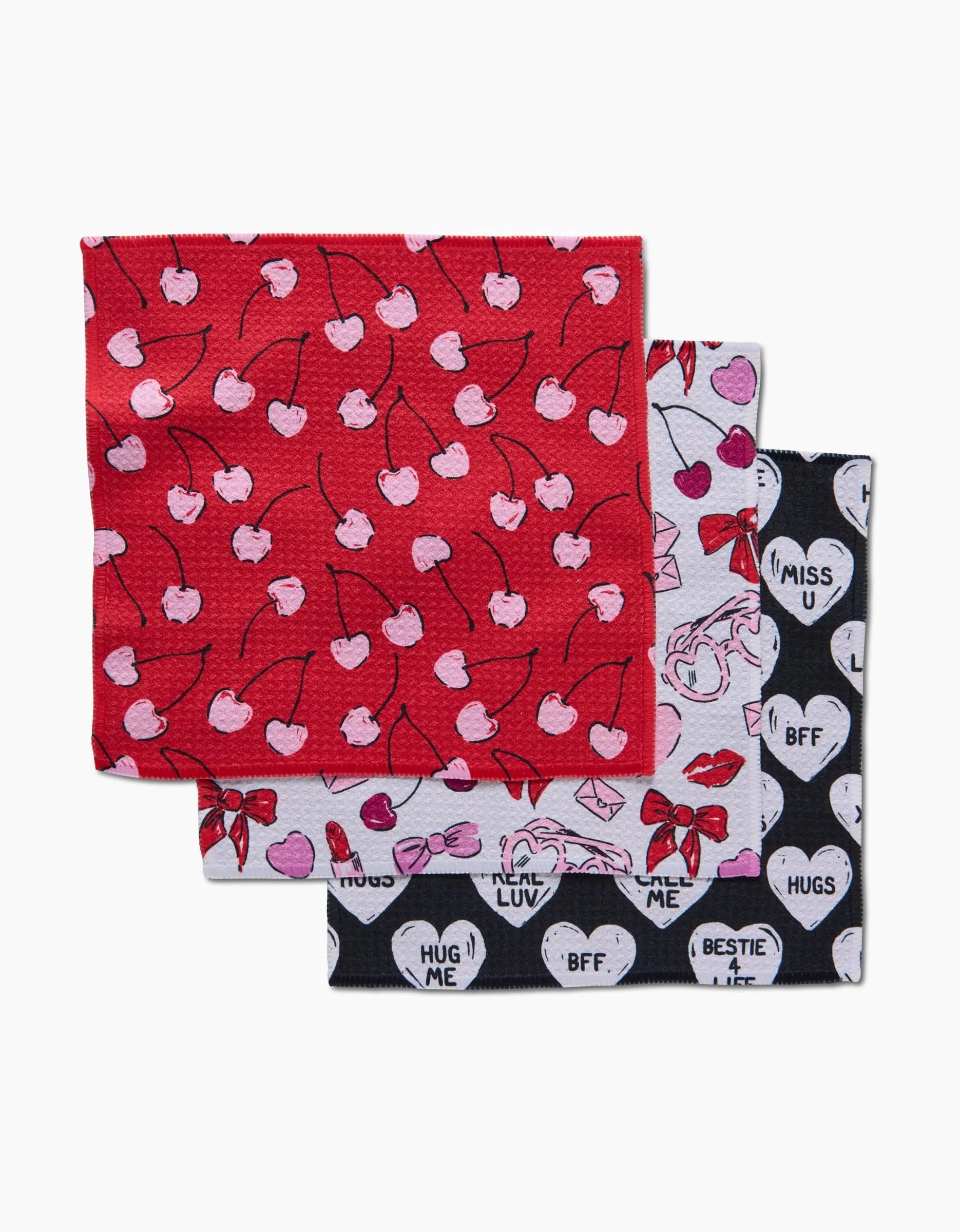 Three folded patterned napkins from the Hufton Studio Valentines dishcloth set: red with pink cherries on top, white with pink bows and hearts in the middle, and black with white hearts and phrases like “I MISS U” and “BFF” on the bottom.