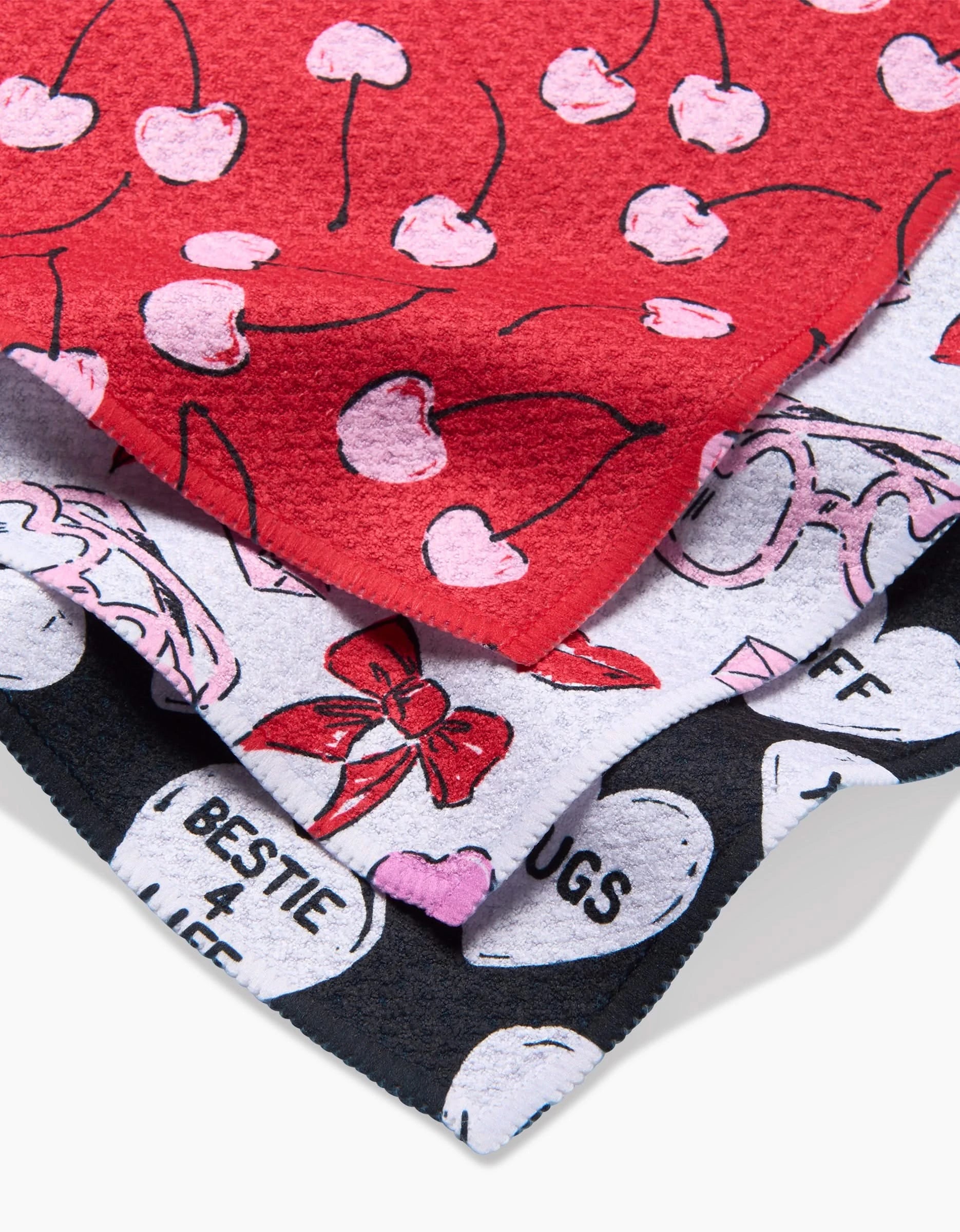 The Valentines towel set by Hufton Studio features three colorful, recycled-material towels: red with pink cherries, white with pink bows and sunglasses, and black with white conversation hearts and friendship-themed words.