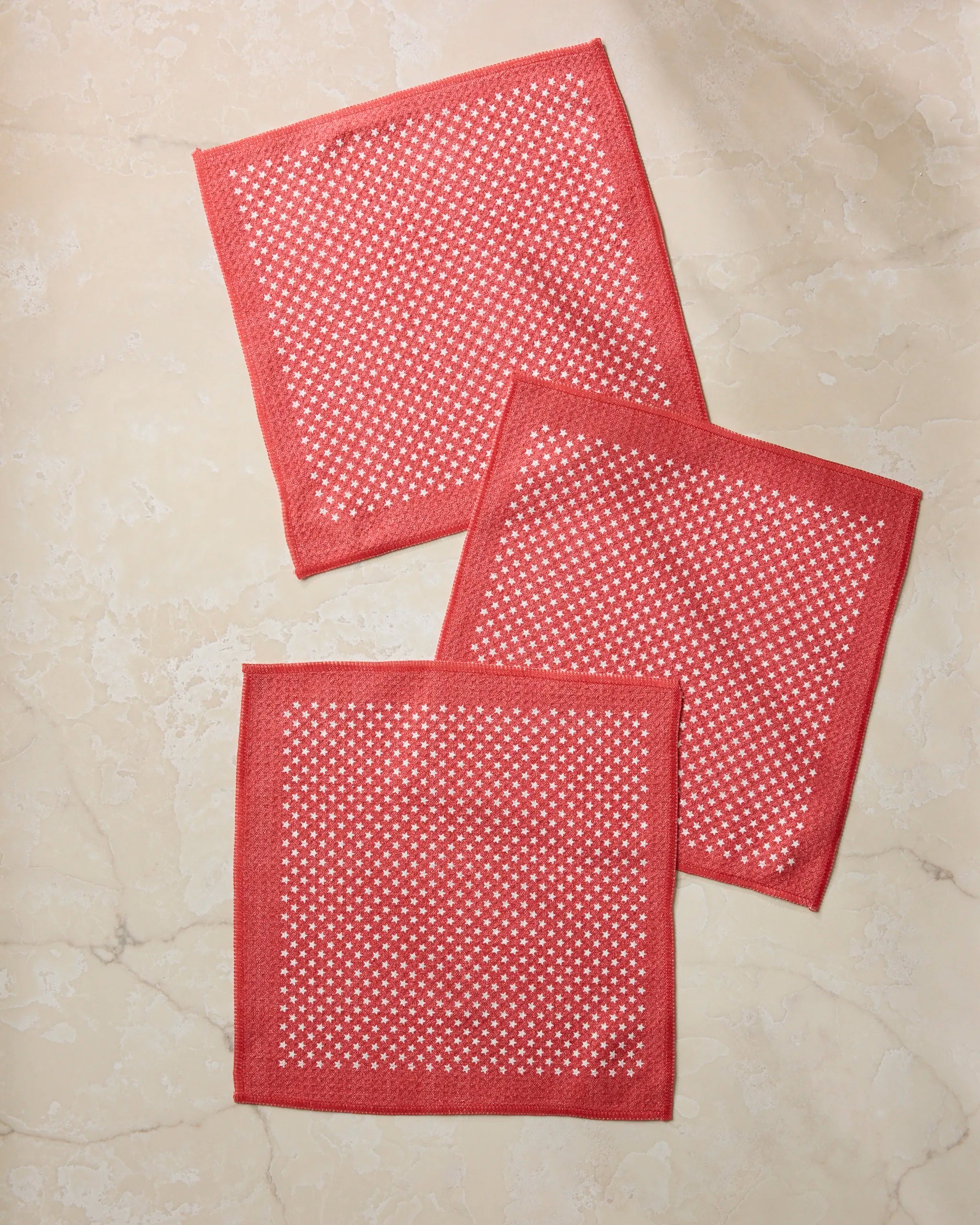 Three Valor napkins by Geometry, featuring a red and white checkered design and made from recycled materials, are casually overlapped on a light beige marble surface.