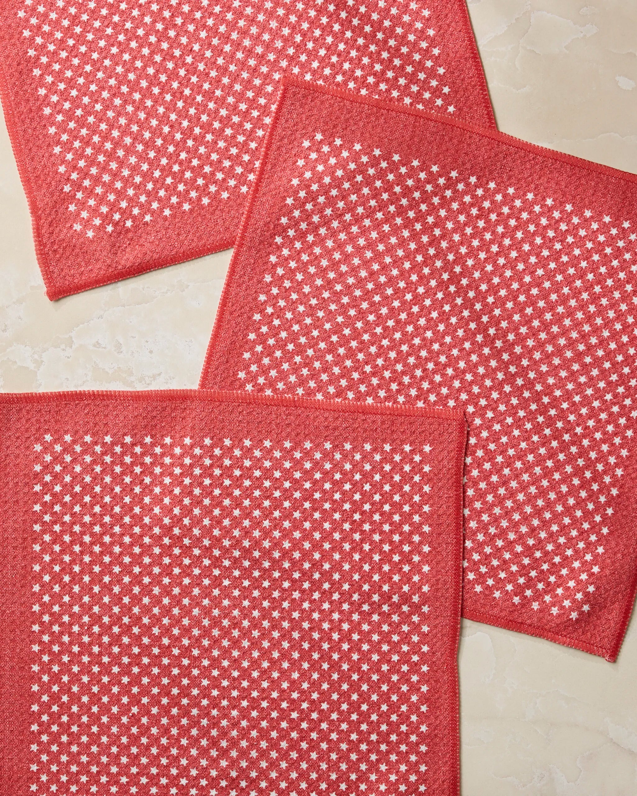 Three Valor cloth napkins by Geometry, featuring a red color with a white star pattern and made from recycled materials, are displayed slightly overlapping on a light, marble-like surface.