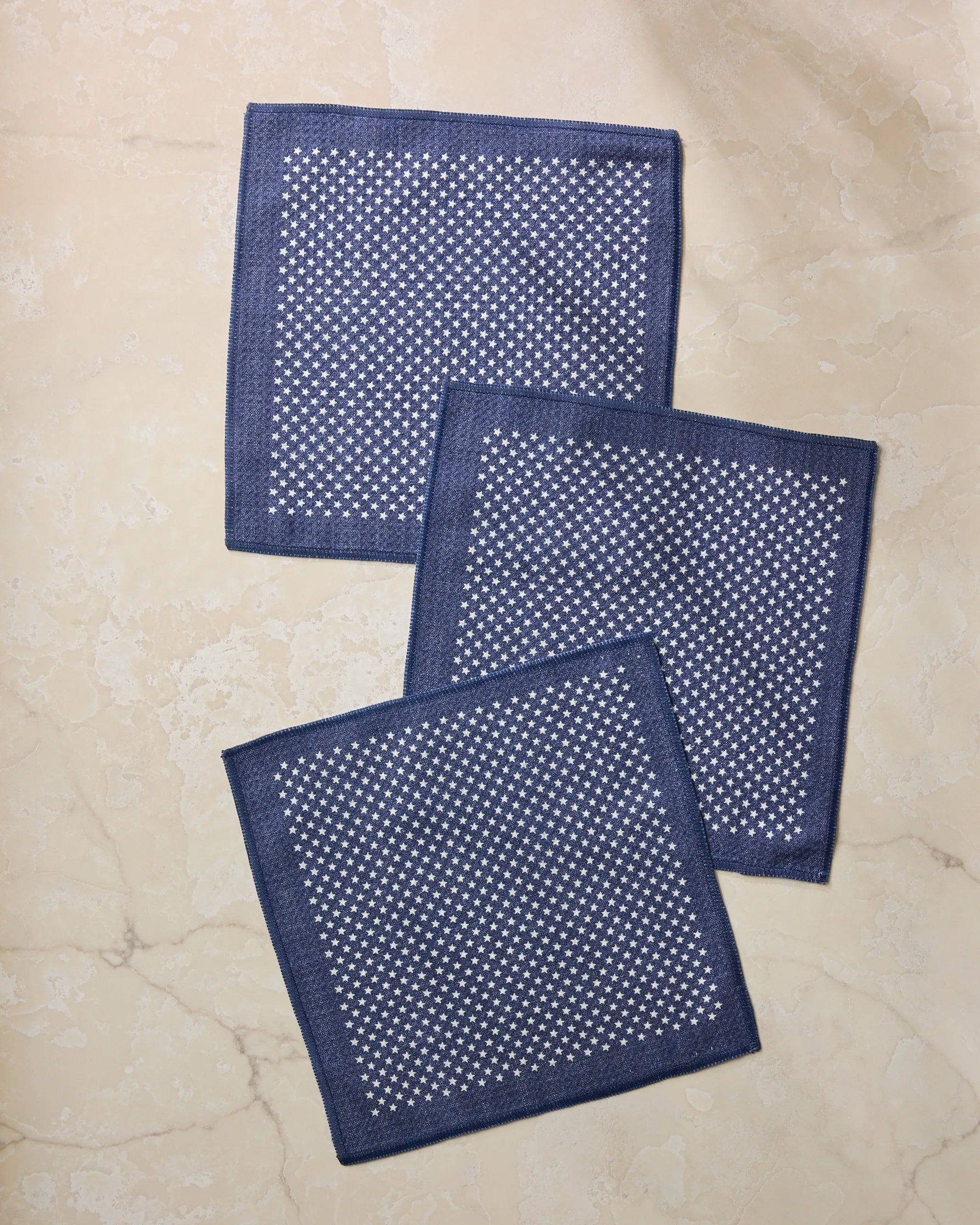 Three Valor navy blue square cloth napkins by Geometry, featuring a white dotted pattern, are displayed overlapping on a light beige marble surface.