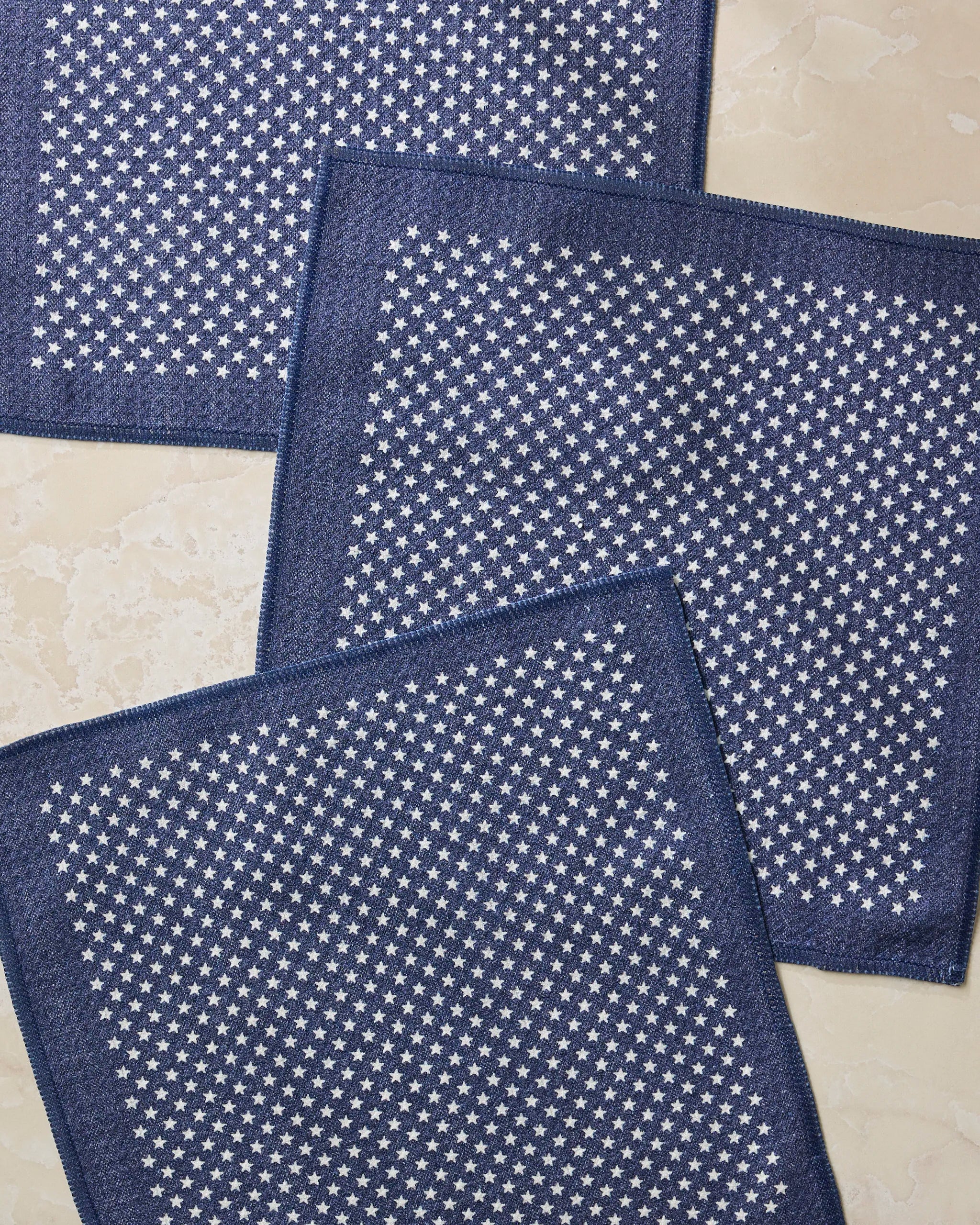 Three Valor navy blue fabric napkins with small white stars, from Geometry’s super-absorbent set made of recycled materials, are arranged on a light beige marble surface.