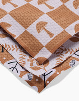 A close-up of three folded Vines Acorns And Mushrooms kitchen towels by Charlie Rowan Designs, made from recycled materials with brown and white patterns featuring mushrooms, leaves, and abstract shapes.