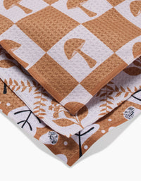 A close-up of three folded Vines Acorns And Mushrooms kitchen towels by Charlie Rowan Designs, made from recycled materials with brown and white patterns featuring mushrooms, leaves, and abstract shapes.