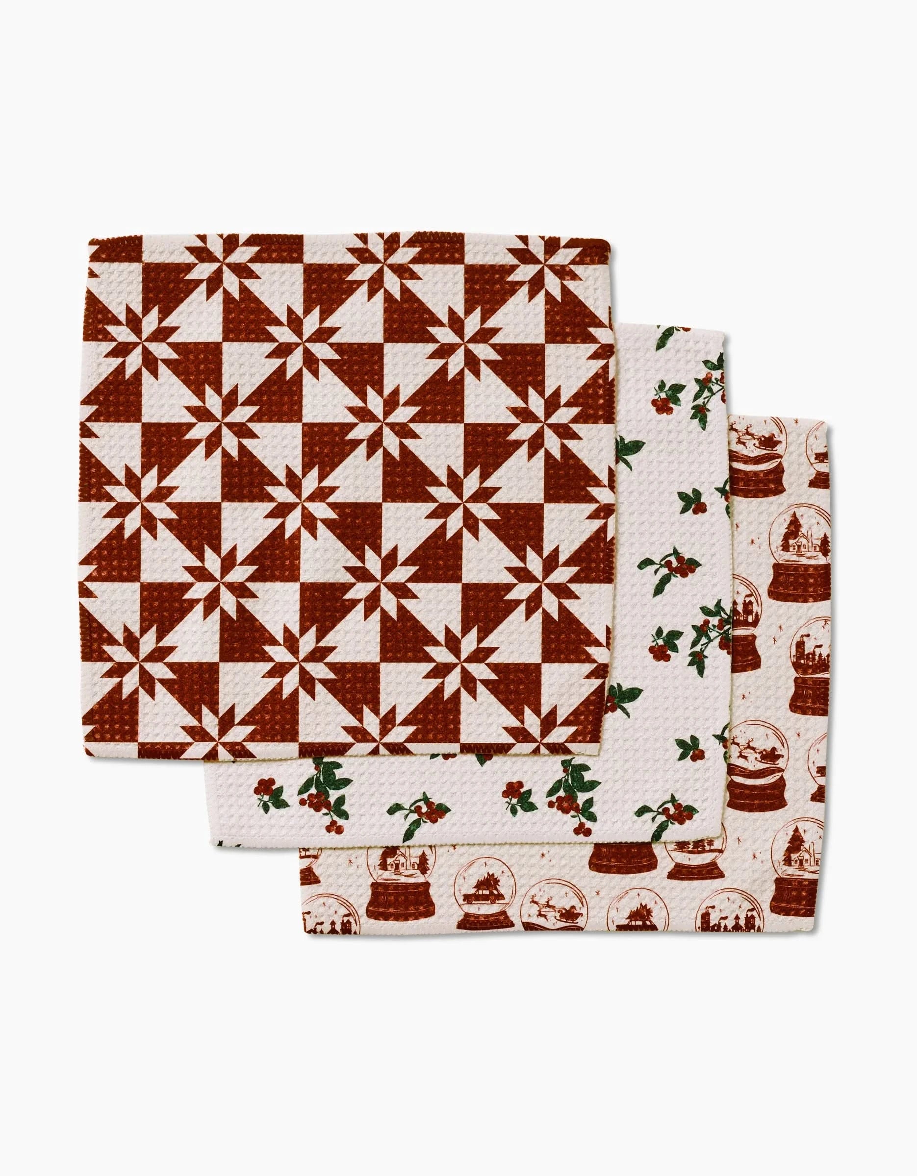 Three Aubrey Fairchild Vintage Christmas Geoweave dishcloths are stacked: the top with a red quilt star design, the middle with holly and berry sprigs, and the bottom featuring red snow globes and holiday scenes on white.