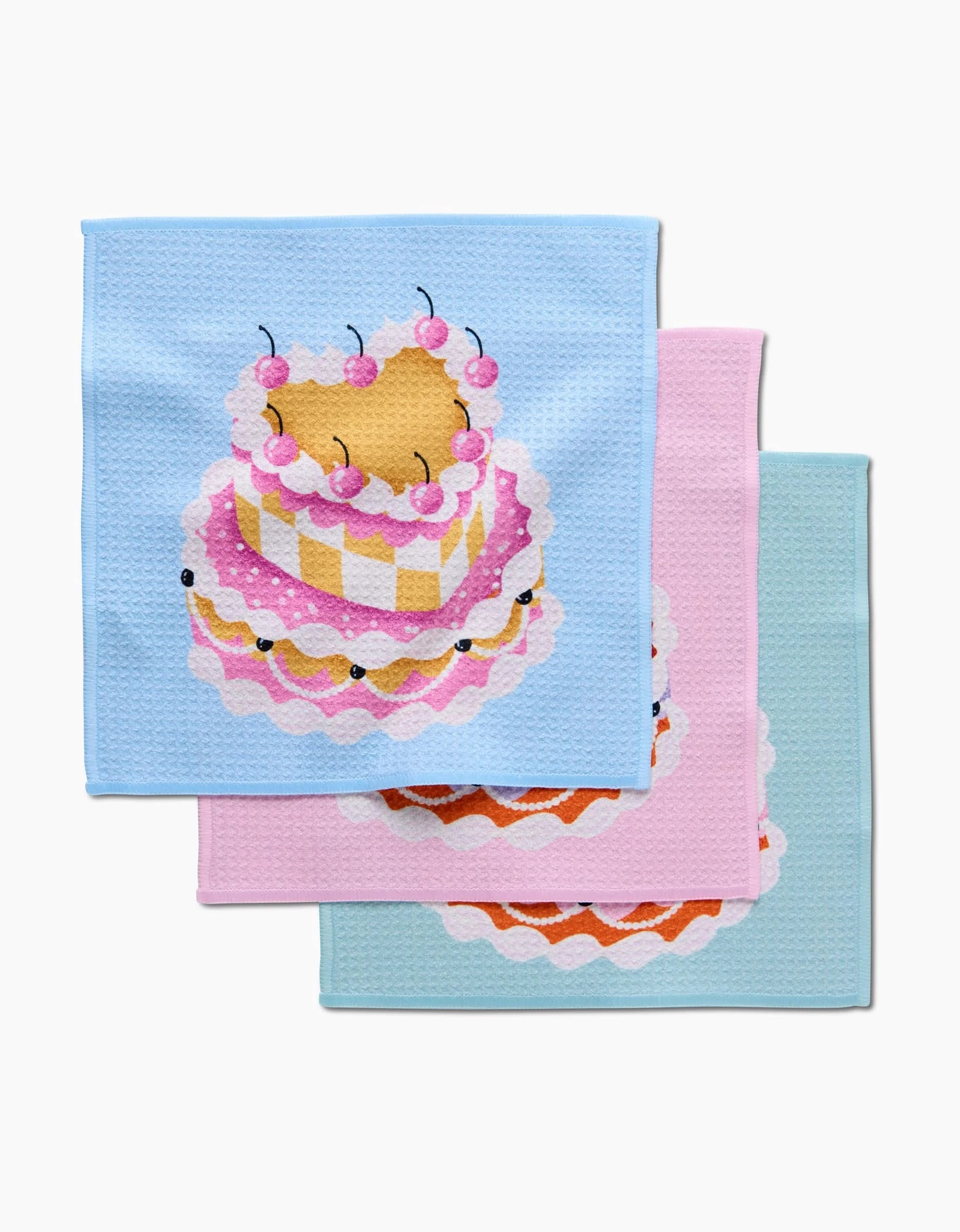The Megan Roy Vintage Heart Cake dishcloth set includes three colorful cloths—light blue, pink, and pale blue—each featuring a whimsical layered cake with cherries. Made from recycled materials and stacked with overlapping corners.