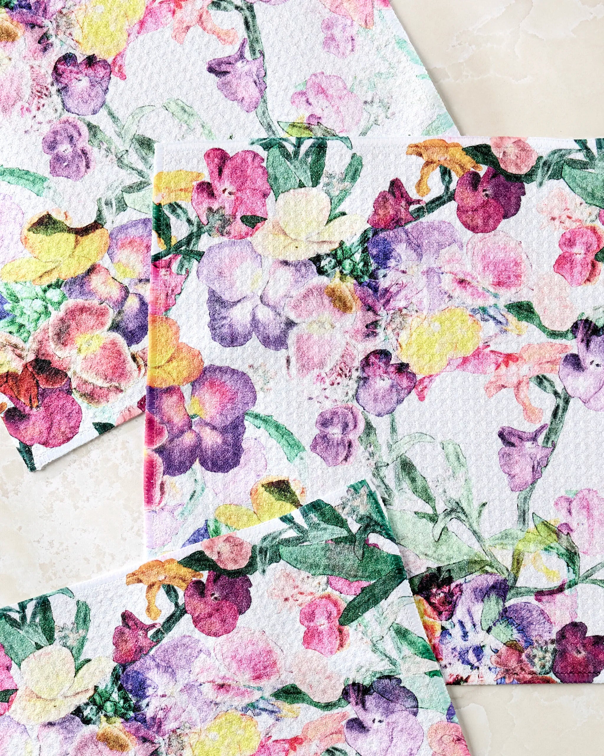 Three Wallflower paper napkins by Pascale Dilger, made from recycled materials and featuring colorful floral patterns with green leaves, overlap on a light, marble-like surface.