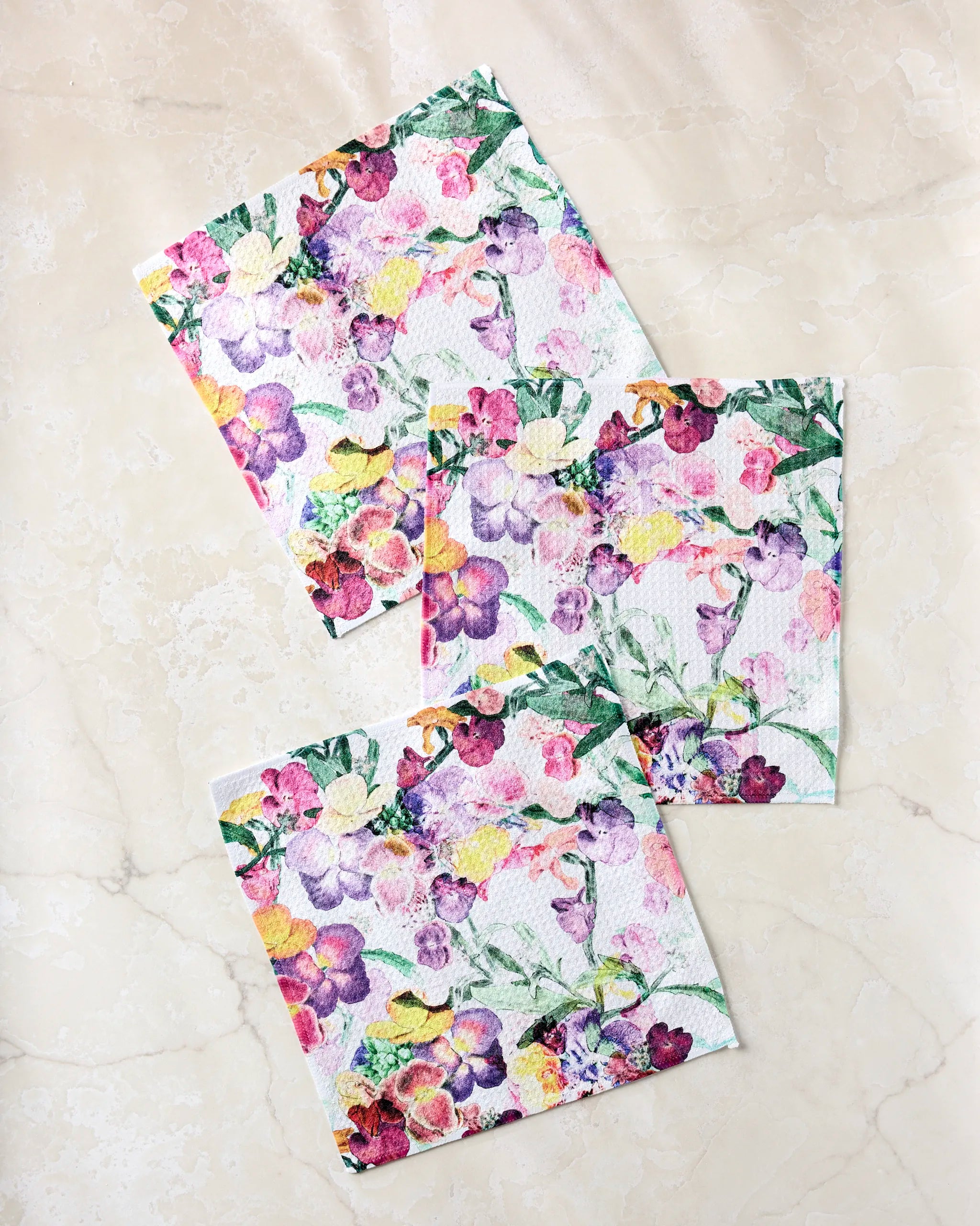 Three Pascale Dilger “Wallflower” square paper napkins, made from recycled materials and featuring a colorful purple, yellow, and pink floral pattern, are arranged on a light marble surface.