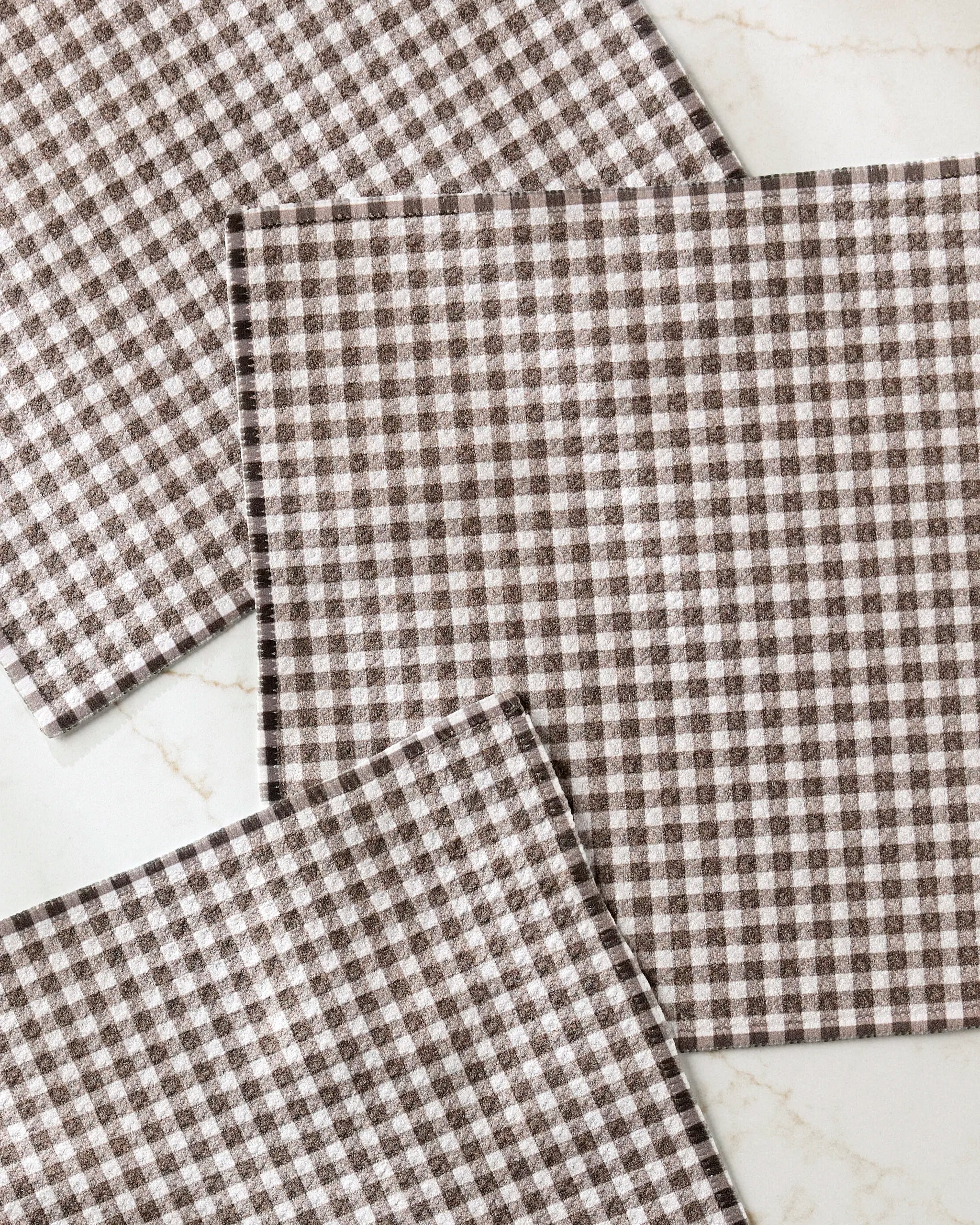 Three Whidbey Check kitchen towels by Jenny Keller, featuring a brown and white checkered pattern, are arranged on a light marble surface. Two towels are angled, partially overlapping the large square napkin in the center.