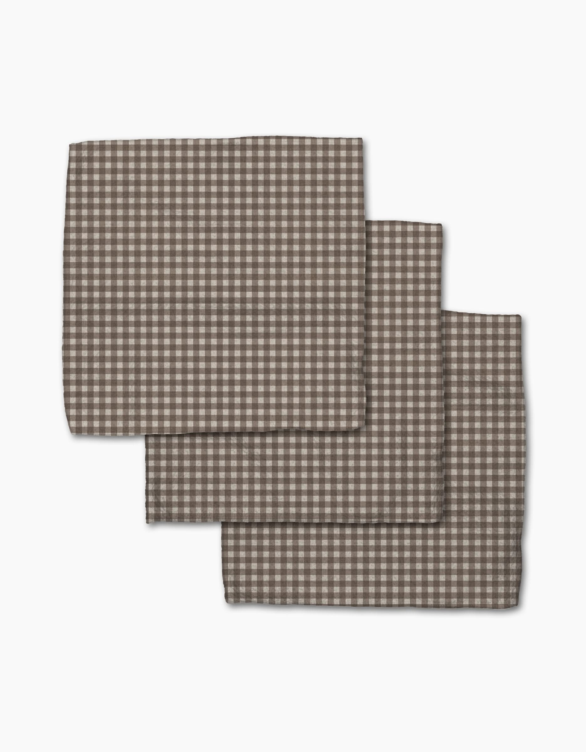 Three Whidbey Check napkins by Jenny Keller, featuring a brown and white checkered design and made from recycled materials, are stacked slightly overlapping on a white background.