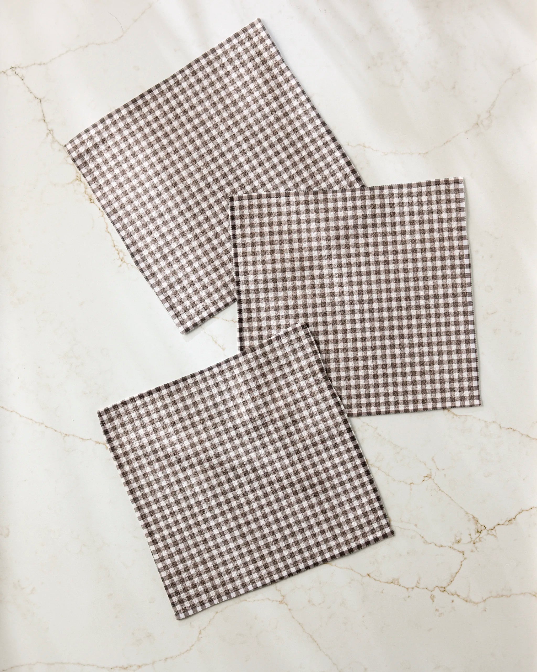 Three Whidbey Check recycled dishcloths by Jenny Keller, featuring a brown and white check pattern, are neatly arranged and slightly overlapping on a light marble surface in a casual, diagonal layout.