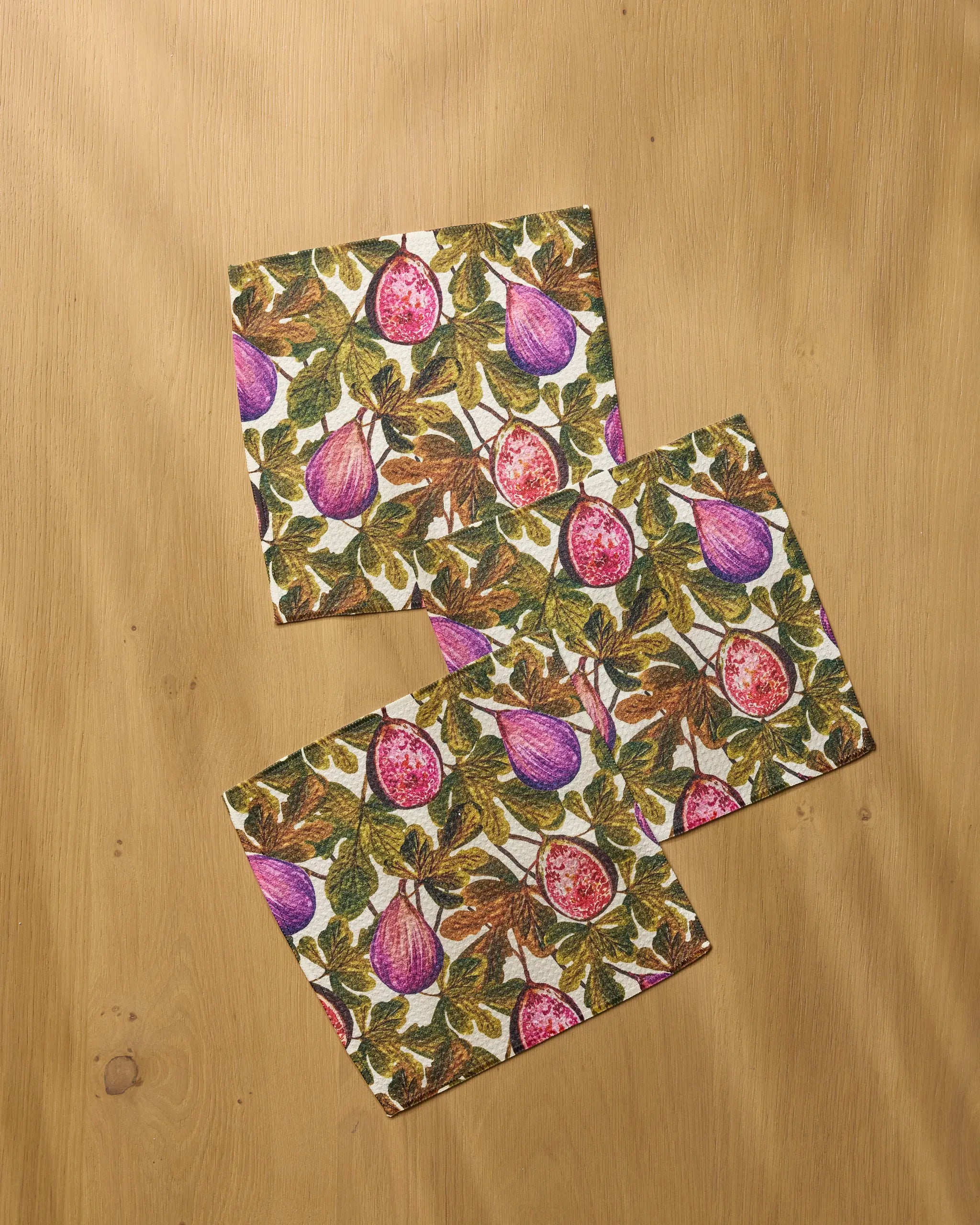 Three Wild Fig napkins by Geometry, featuring a vibrant purple fig and green leaf pattern, are arranged on a light wooden surface. Made from recycled materials, these eco-friendly napkins add a natural touch to your table setting.