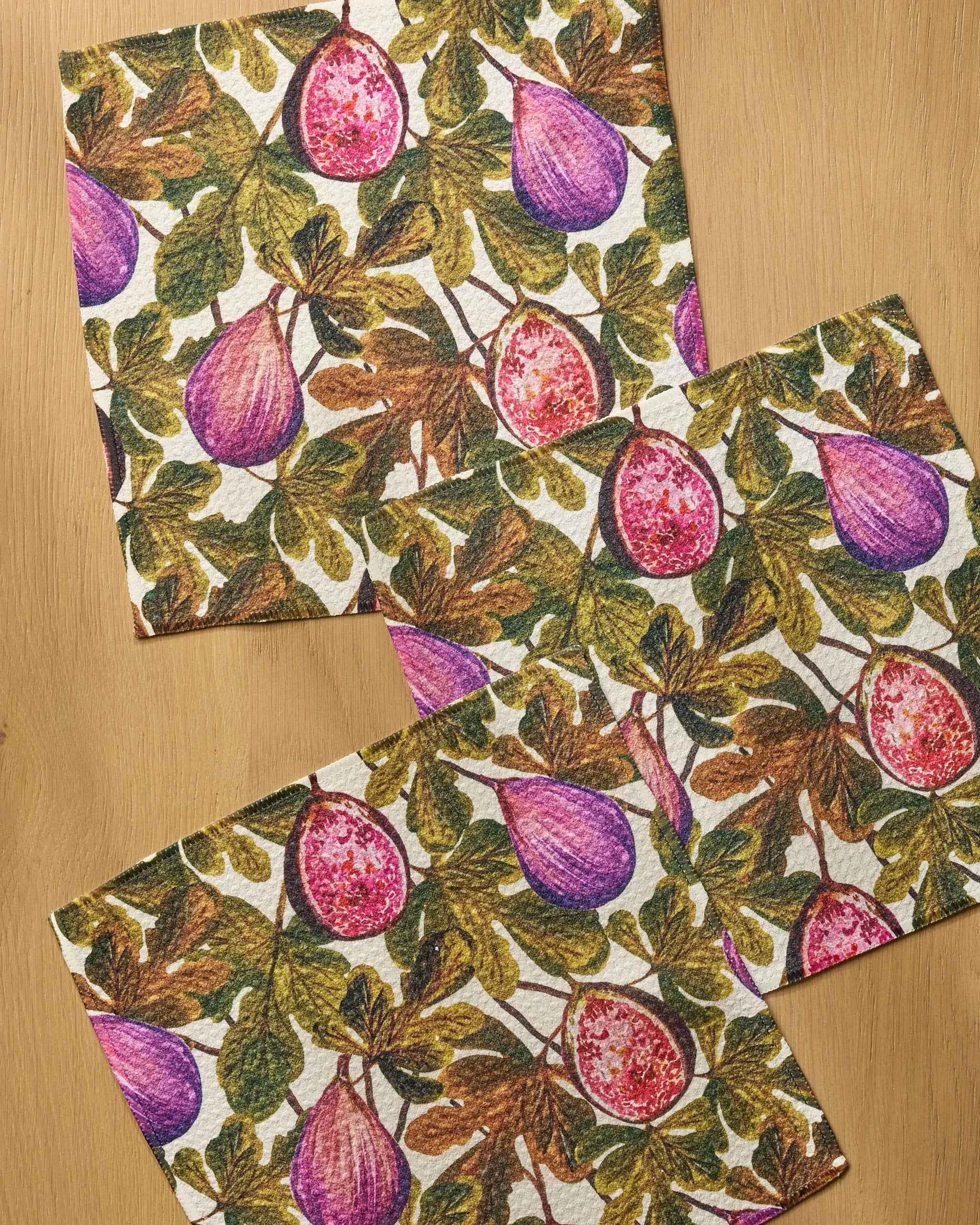 Three super-absorbent Geometry Wild Fig dishtowels featuring a purple, pink, green, and brown fig-and-leaf pattern are arranged on a light wooden surface.