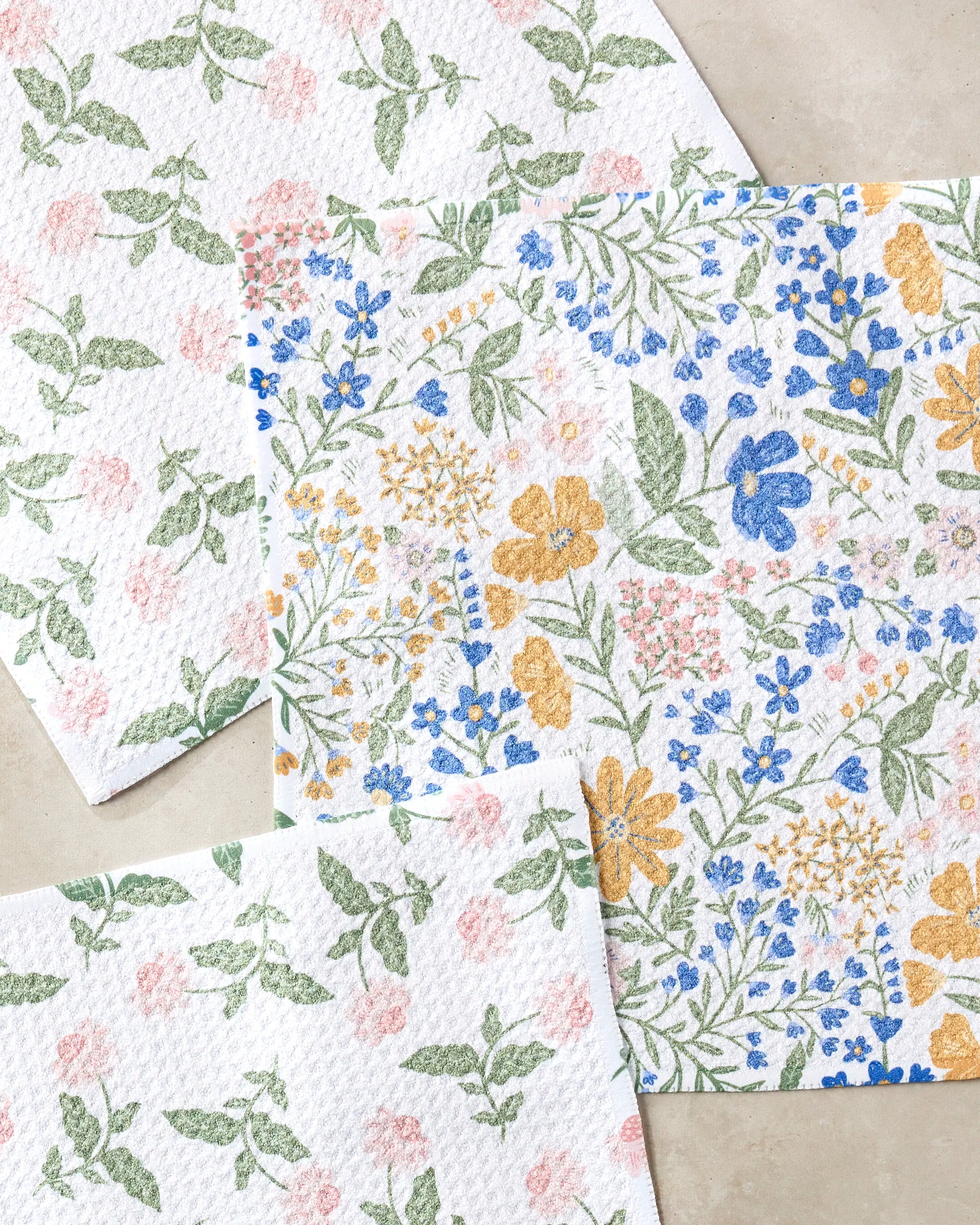 Three Wildflower Vine Blush dishtowels by Danelys Sidron, featuring colorful blue, yellow, and pink floral patterns with green leaves, are displayed on a neutral background.