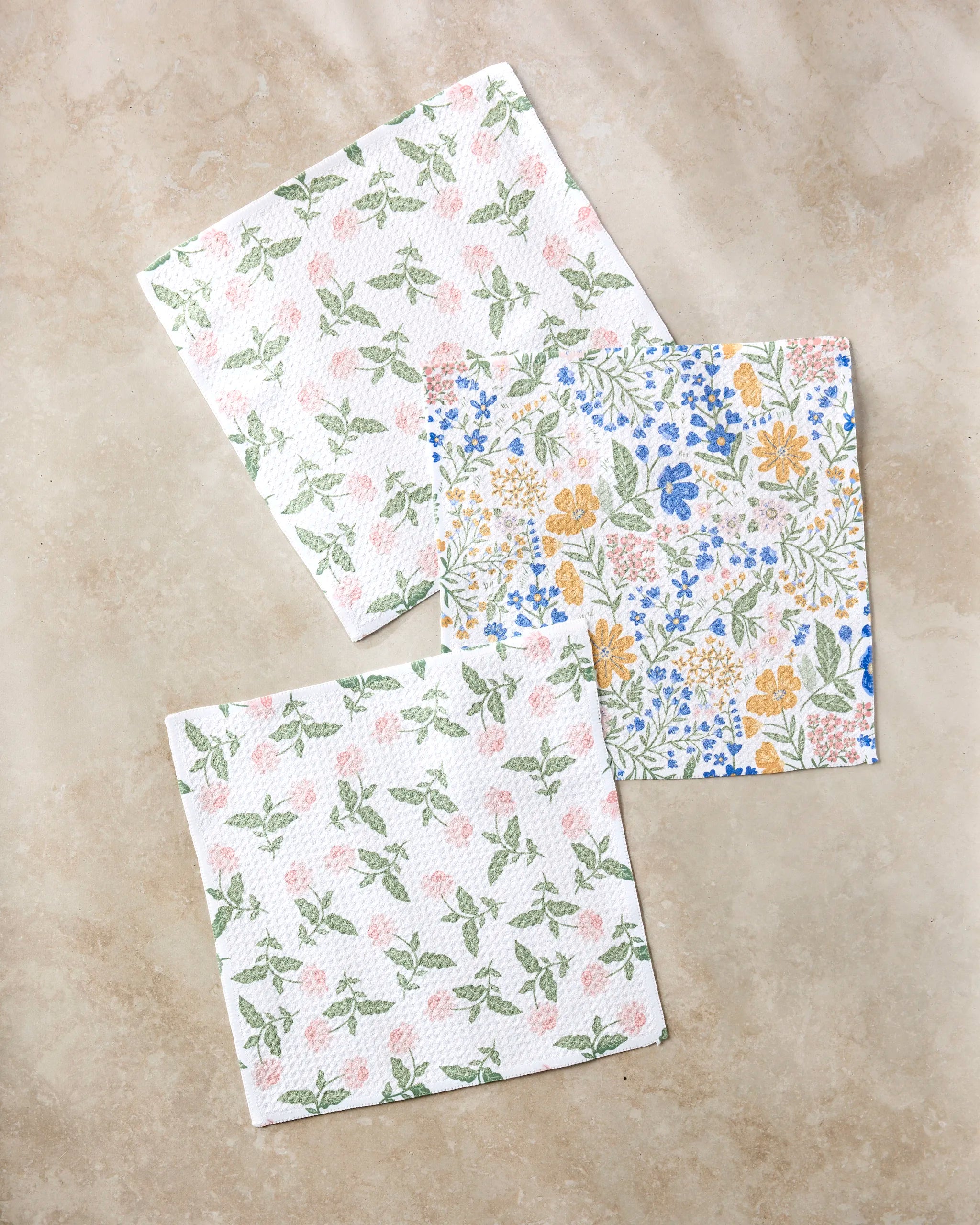 Three Danelys Sidron "Wildflower Vine Blush" paper napkins rest on a beige surface: two with green and pink leaves, one with yellow, blue, and green florals—crafted from recycled materials for an eco-friendly touch.
