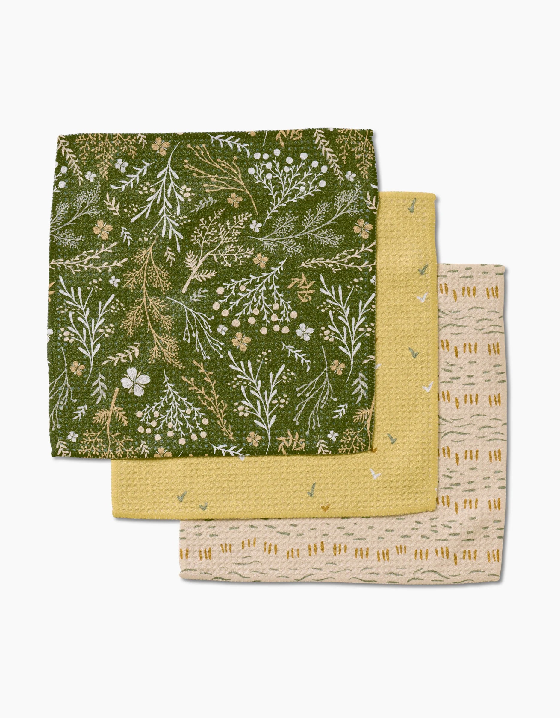 Three folded Wildflowers dishcloths by Danelys Sidron are stacked: the top is green with a white and yellow botanical pattern, the middle is solid yellow with subtle texture, and the bottom is beige with yellow and brown geometric designs.