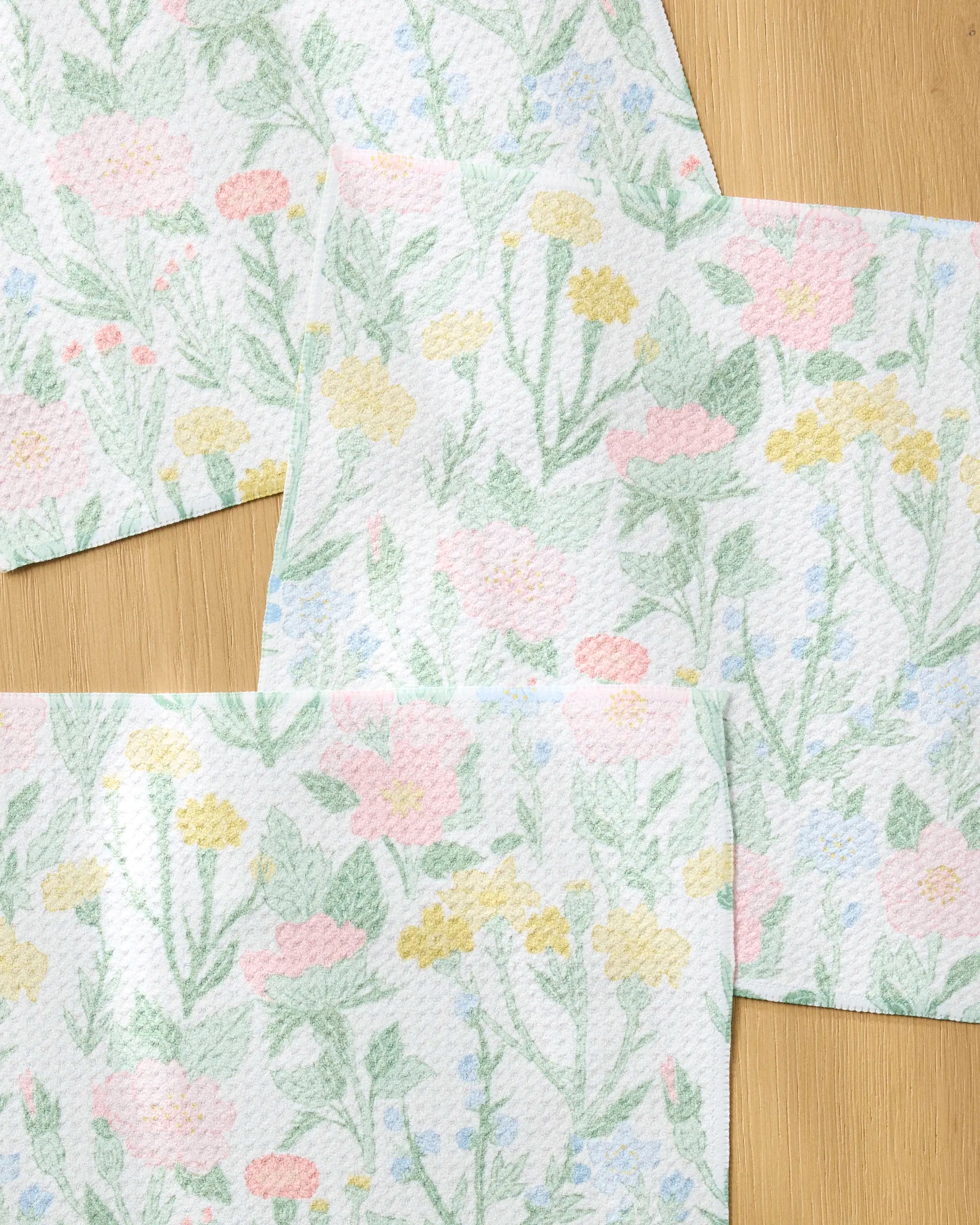 Wildflowers in Bloom paper towels by Jasmine Blooms Designs, made from recycled materials, showcase pastel pink, yellow, and green flowers and leaves overlapping on a light wood surface.