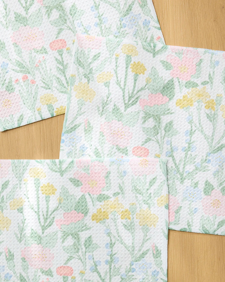 Wildflowers in Bloom paper towels by Jasmine Blooms Designs, made from recycled materials, showcase pastel pink, yellow, and green flowers and leaves overlapping on a light wood surface.