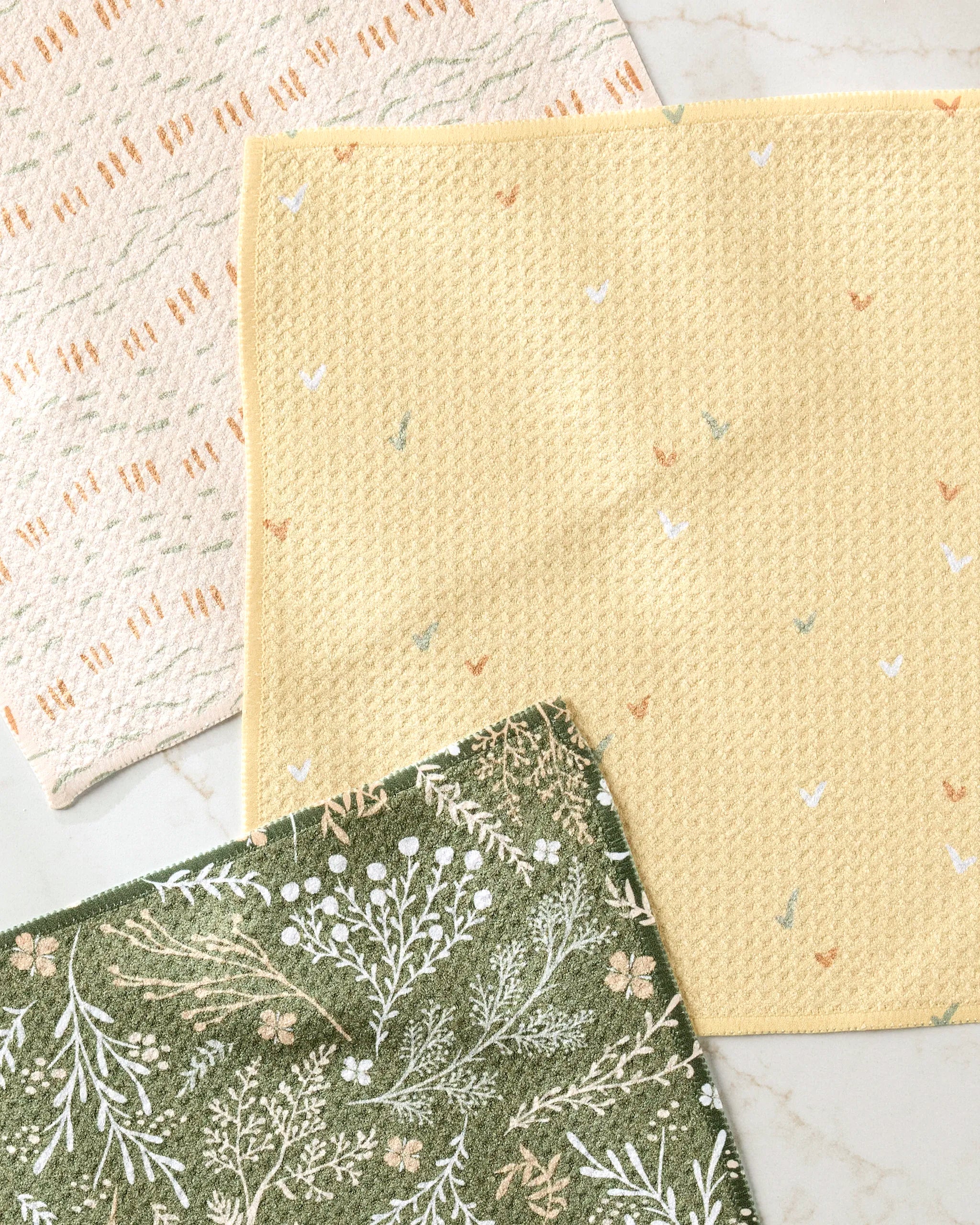 Three cloths from Danelys Sidron’s Wildflowers set overlap on marble: pale yellow with white and orange, sage green with white florals, and cream with orange shapes—a charming, fast-drying dishtowel trio.