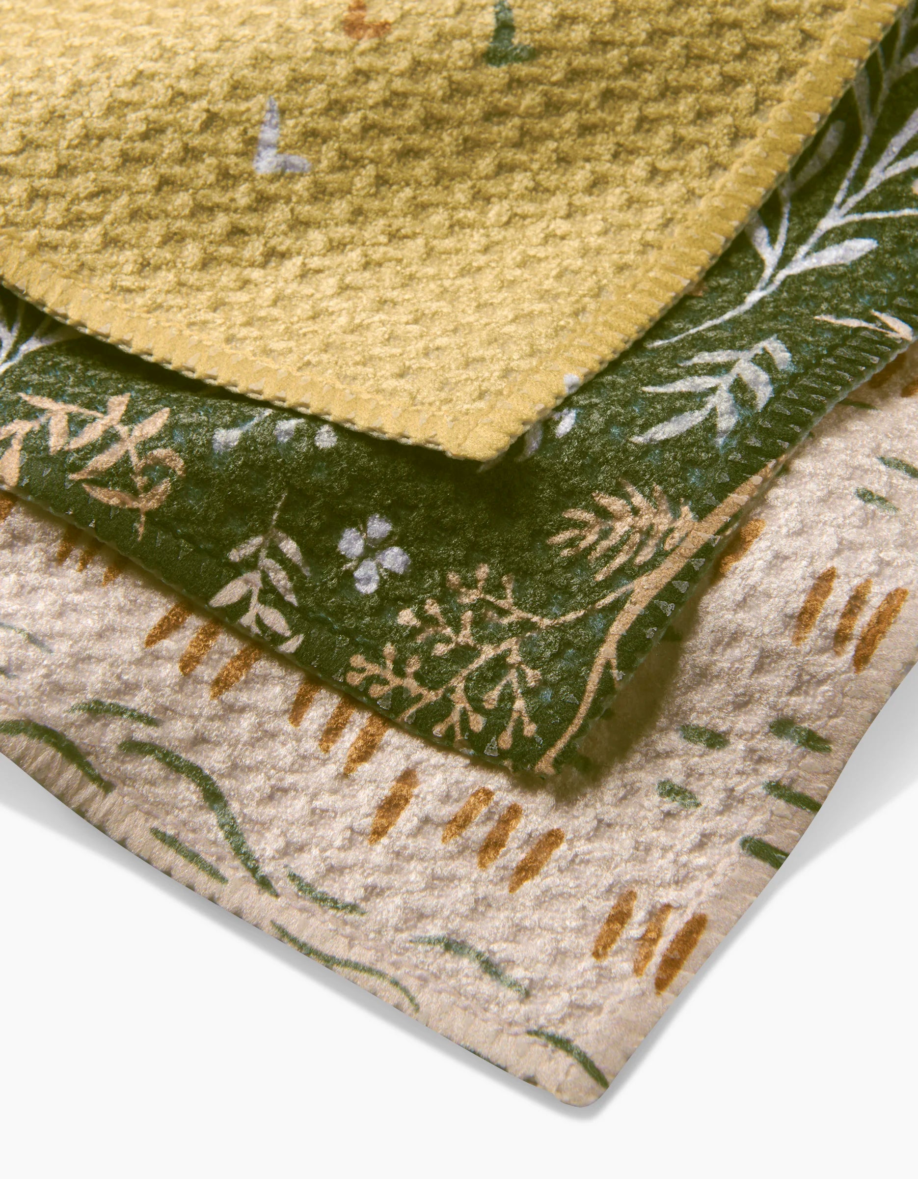 Three folded Danelys Sidron Wildflowers blankets: the top is yellow Geoweave with a waffle pattern, the middle is green with white and yellow botanical designs, and the bottom is cream with green and brown abstract lines.