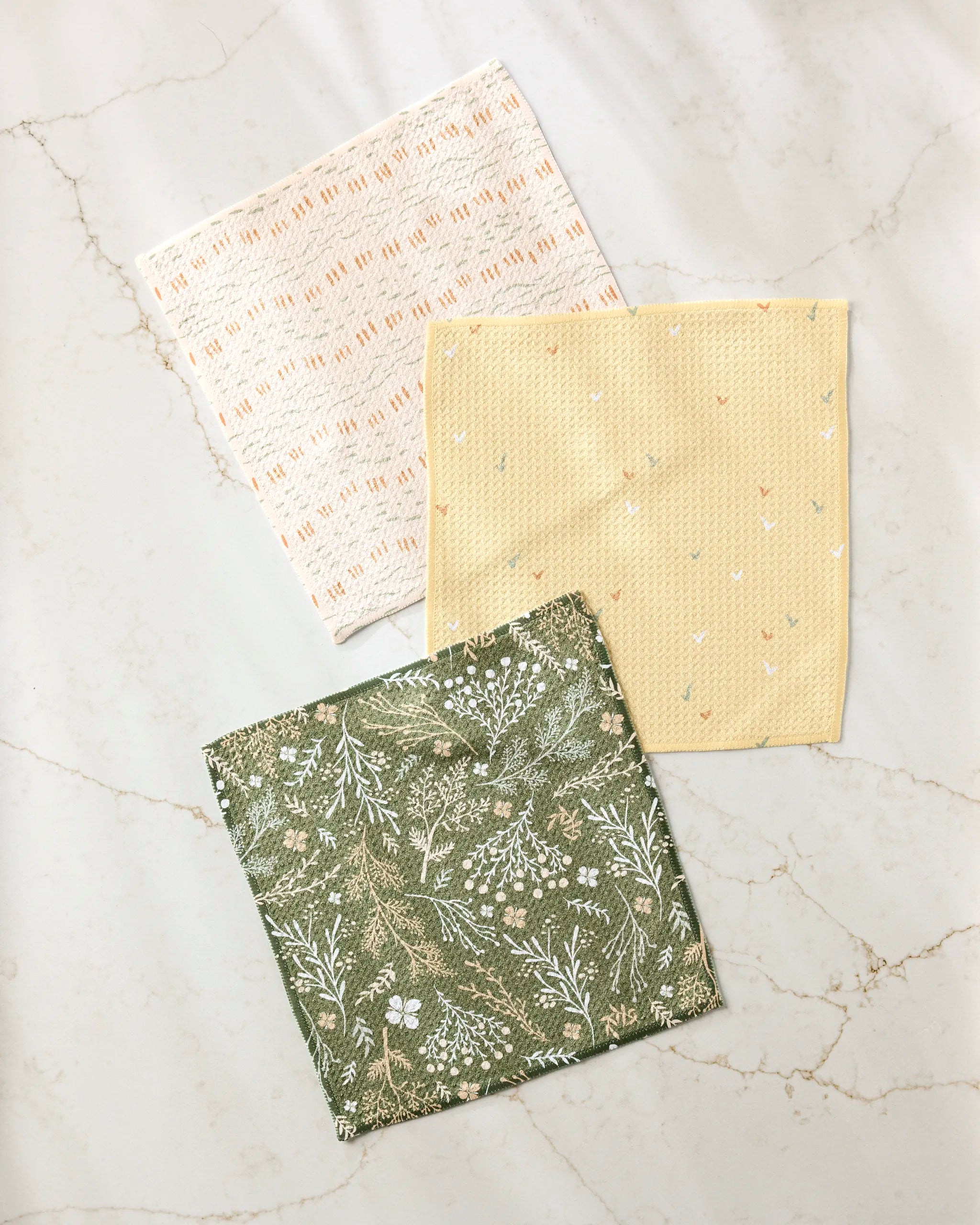 Three reusable Danelys Sidron Wildflowers cloths, including a cream one with an orange geometric pattern, a yellow one with small shapes, and a super-absorbent green dish towel, are arranged on a light marble surface.