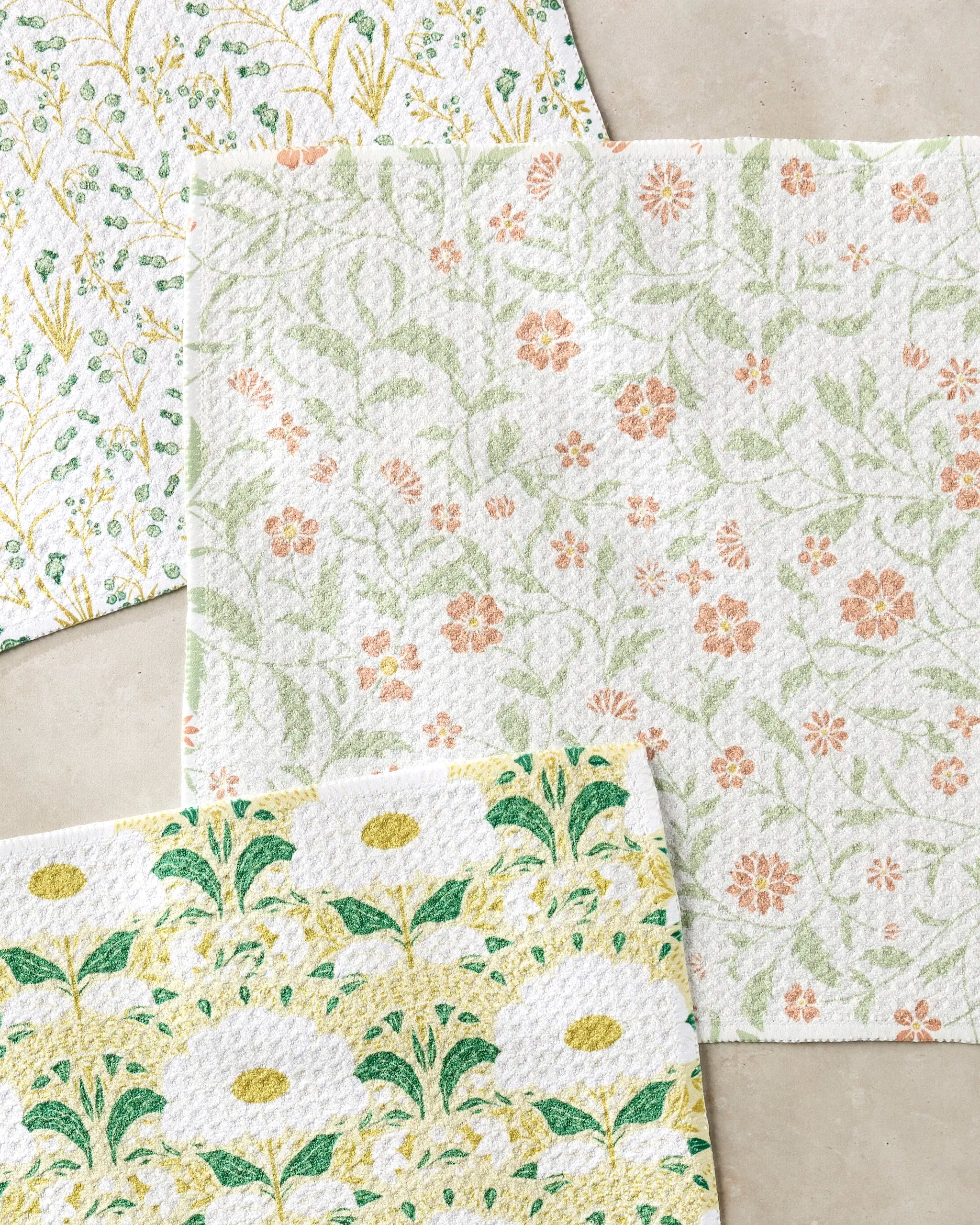 Three Wildlight rectangular recycled dishcloths by Danelys Sidron, in green, yellow, and peach florals, overlap on a beige surface. Each super-absorbent towel features a unique botanical pattern.