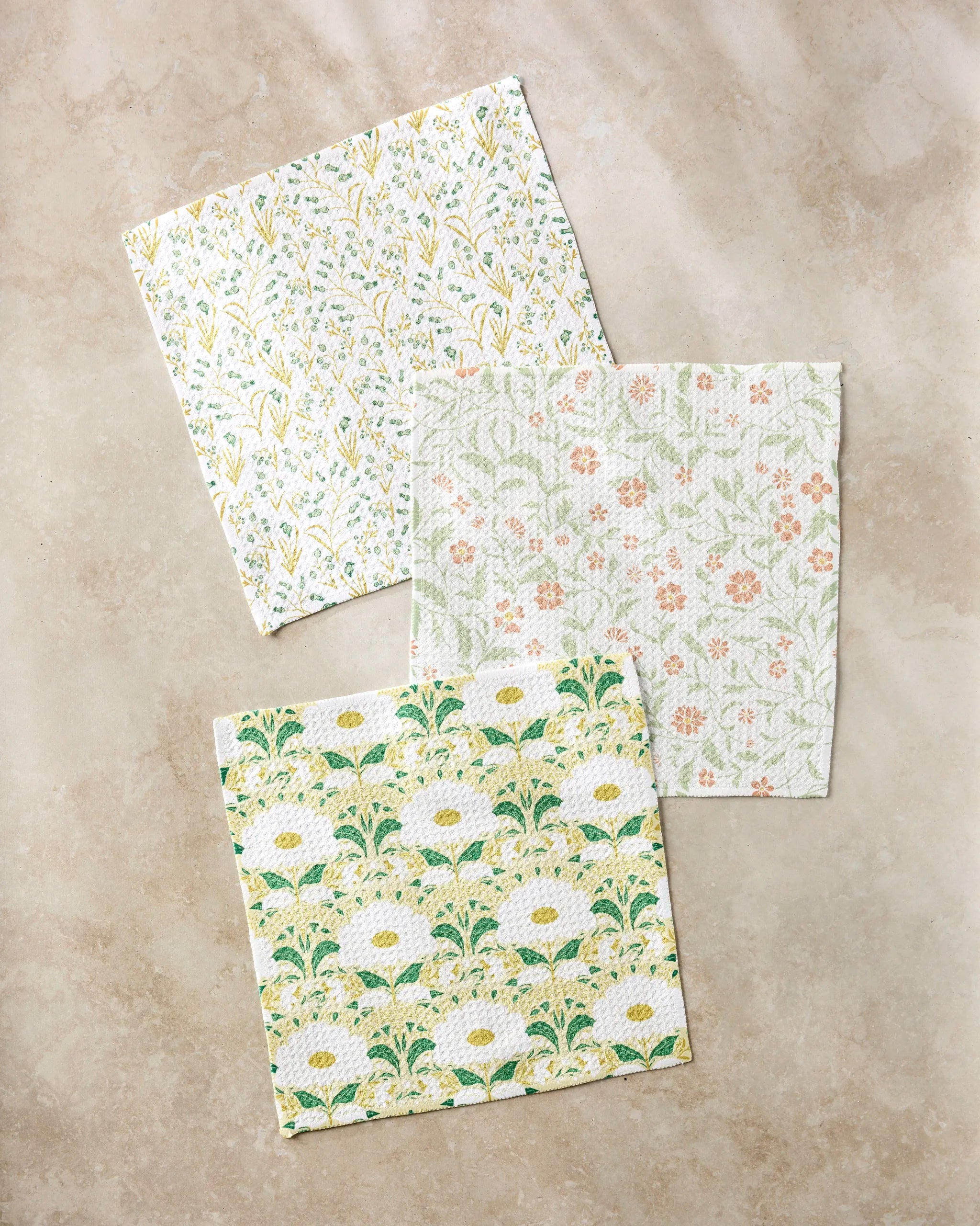 Three Wildlight napkins by Danelys Sidron, crafted from recycled materials, display unique floral patterns in pastel yellow, green, orange, and white. They’re arranged on a beige stone surface.