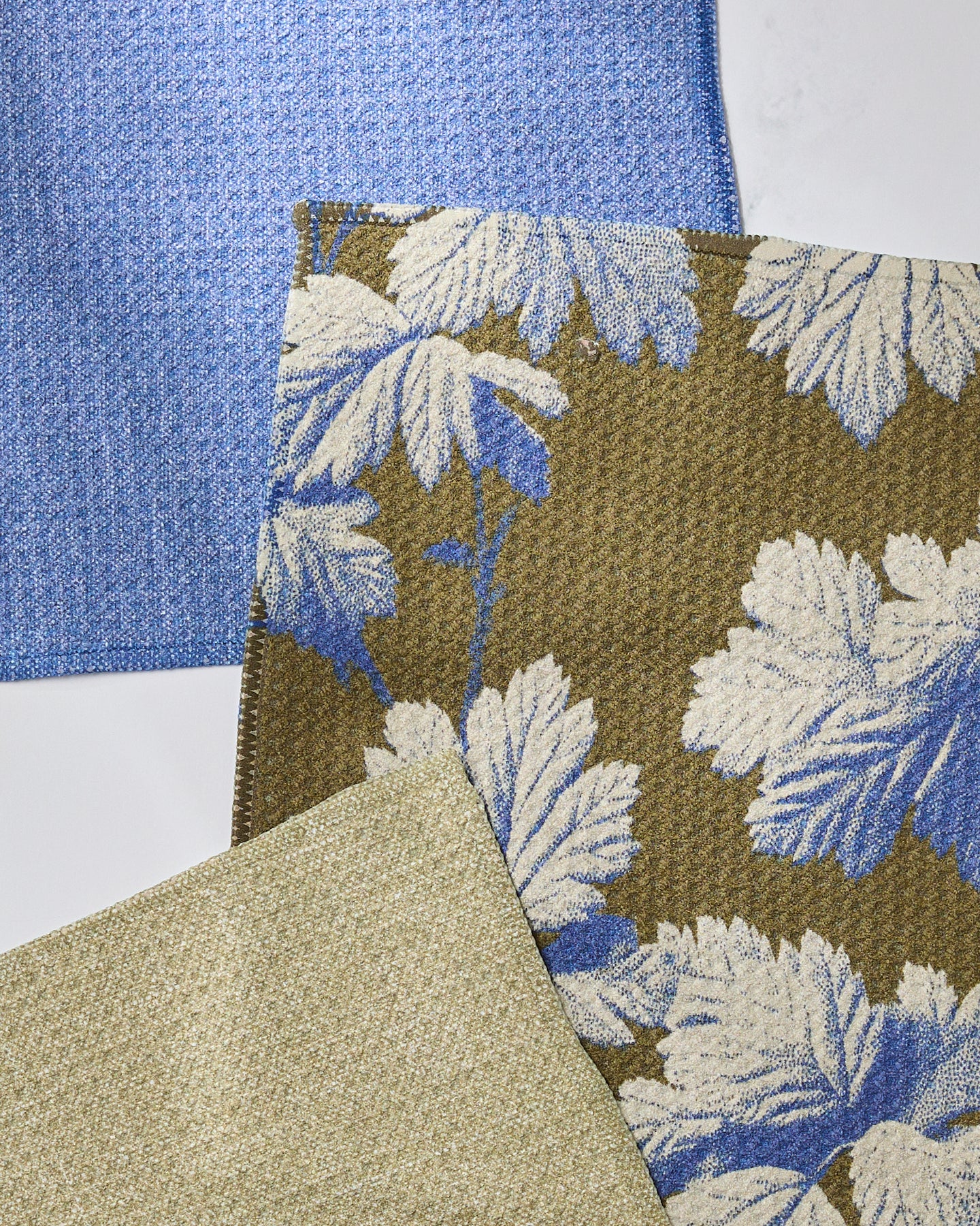 Three overlapping fabric swatches from Geometrys Wildshade: a blue textured, a beige, and a brown with large blue and white leaves—ideal for inspiration when creating super-absorbent dishtowels or dishcloths. Swatches shown on a white background.