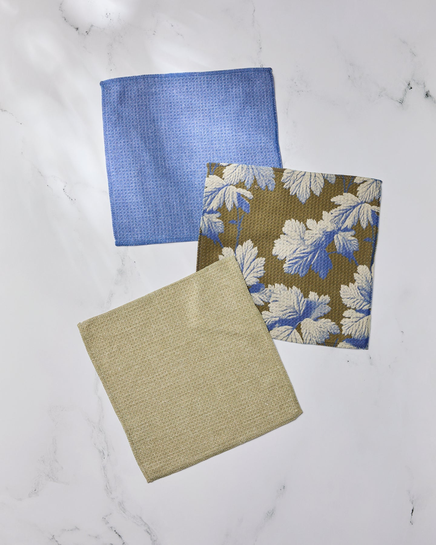 Three folded Geometry Wildshade fabric napkins, made from recycled materials, are displayed on white marble: one blue, one beige, and one brown with a blue and white leaf pattern.