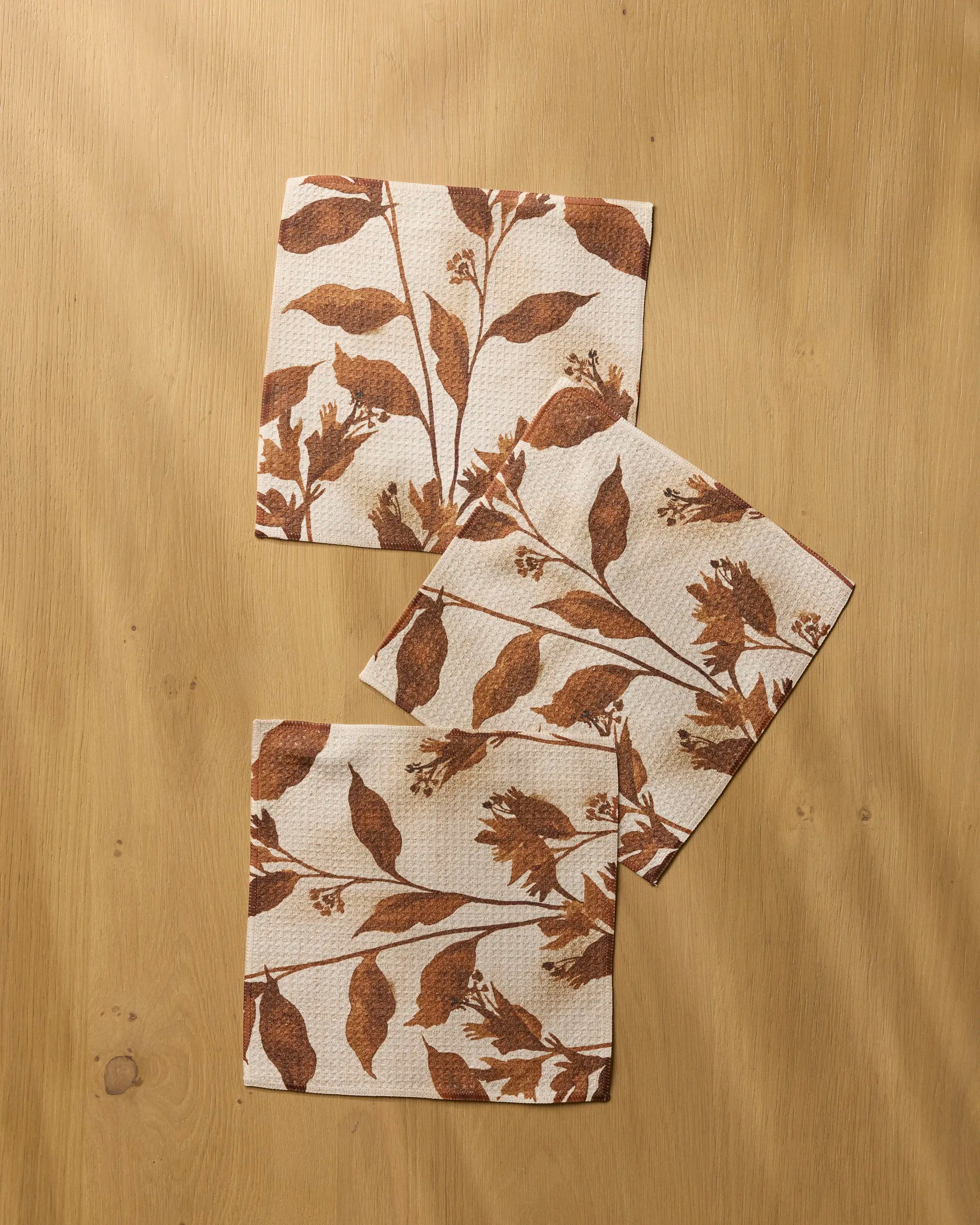 Three Willowmere kitchen towels by Geometry, featuring brown botanical leaf prints, are arranged overlapping on a light wooden surface with soft shadows.