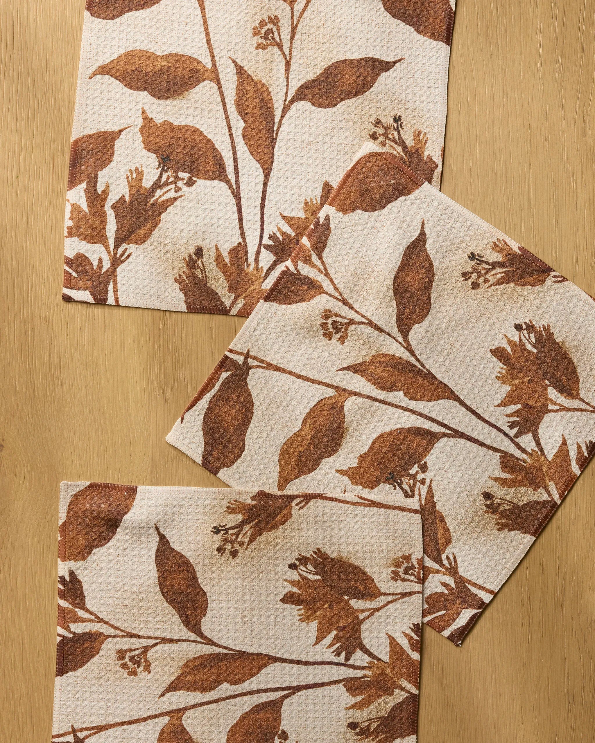 Three Willowmere paper napkins from Geometry, featuring a brown botanical leaf and branch design and a textured look, are slightly overlapping on a wooden surface.