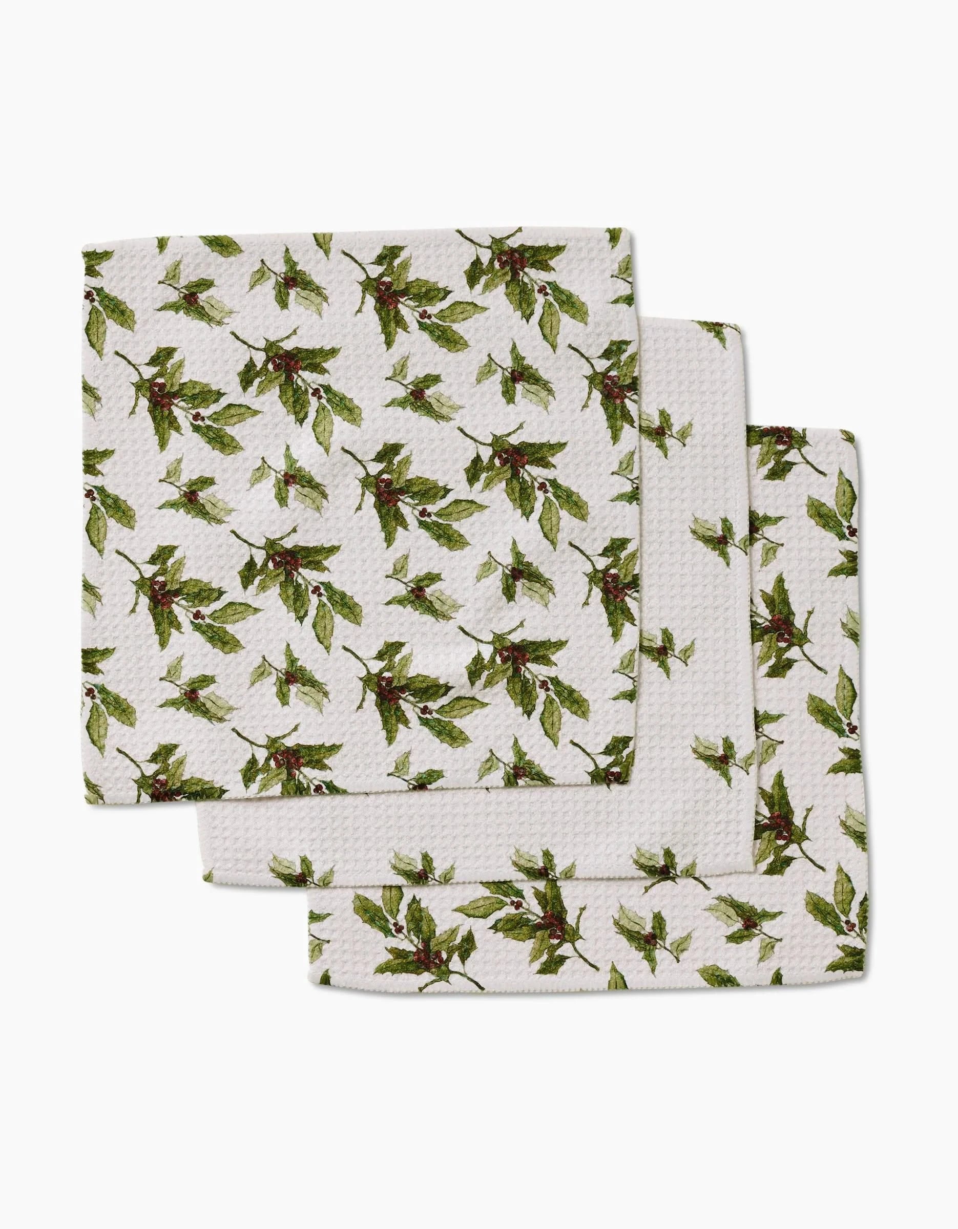 The Aubrey Fairchild Winter Berry set includes three white kitchen towels made from recycled materials, each featuring a green and red leafy botanical print, stacked neatly against a white background.