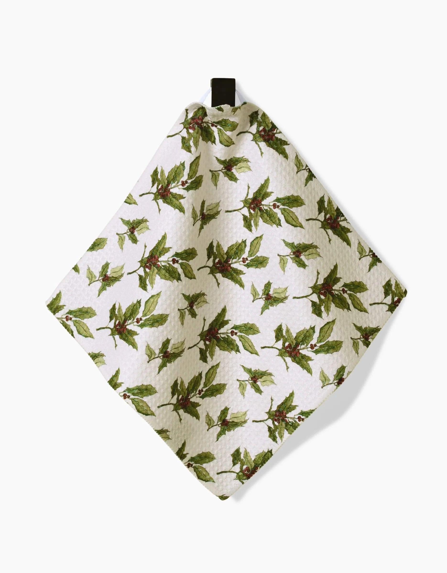 The Aubrey Fairchild Winter Berry dishcloth, made from Geoweave fabric, showcases a white textured design with green leaves and red berries. It hangs by a black top loop against a plain background.