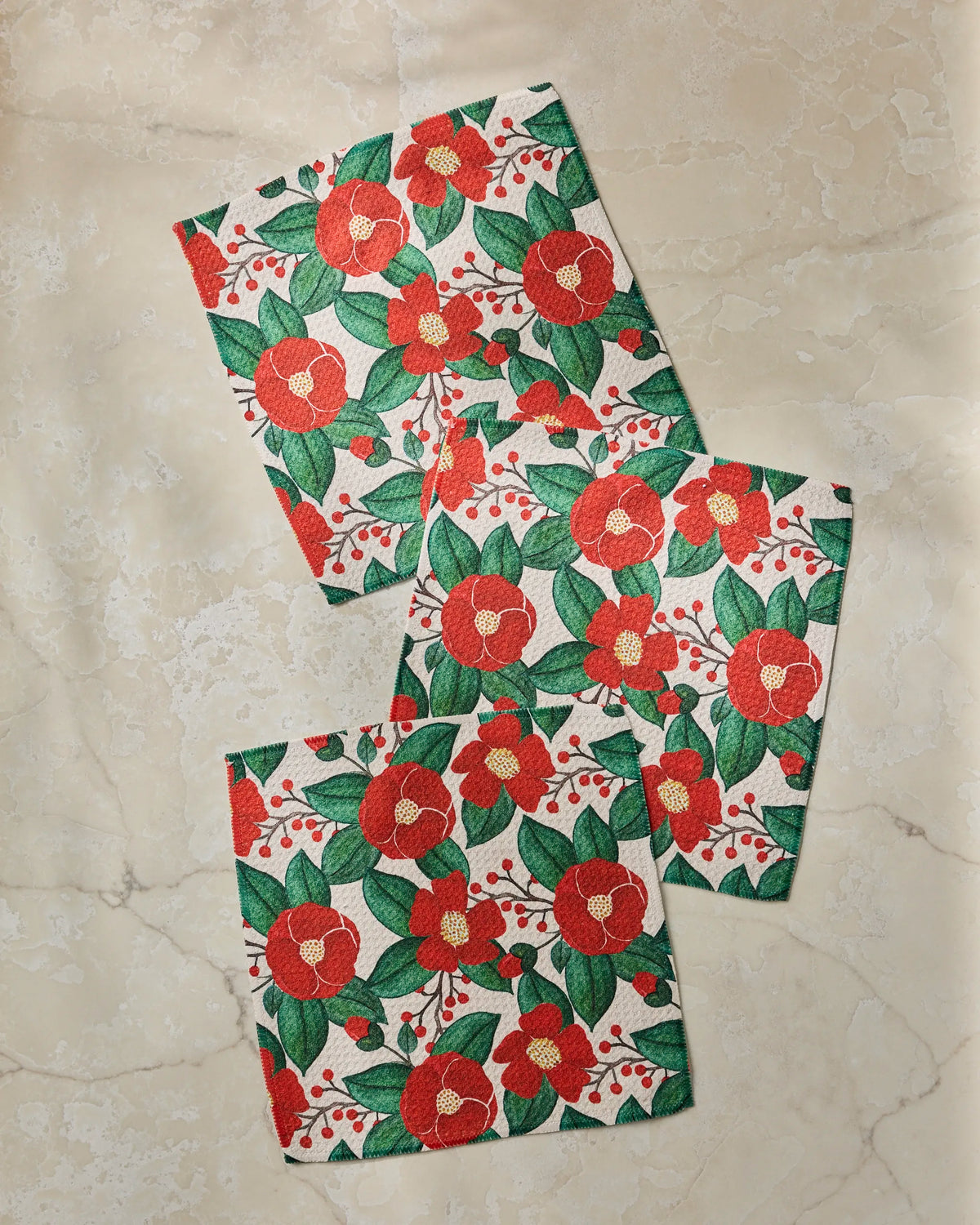 Four rectangular cloth napkins from Geometry’s Winter Camellia collection, crafted from recycled materials and featuring a red and green floral pattern, are arranged on a light marble surface. The flowers display red petals with yellow centers and green leaves, accented by small red berries throughout.