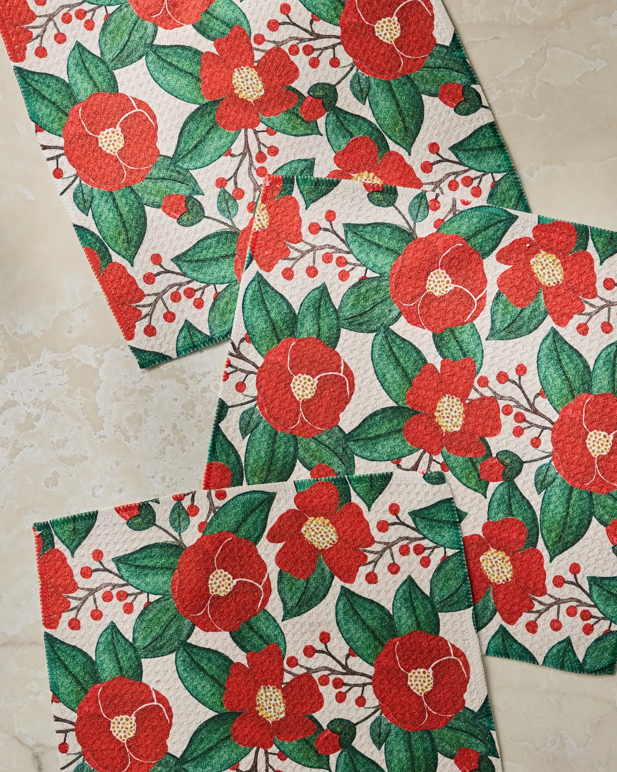 Three festive Winter Camellia napkins by Geometry, featuring red flowers, green leaves, and yellow centers, overlap on a light marble surface. Crafted from recycled materials for an eco-friendly touch, the design showcases eye-catching red berries and intricate textured details.