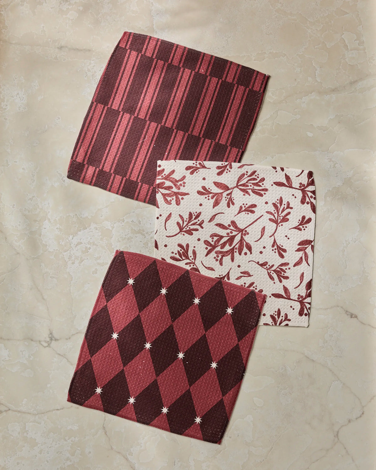 Three napkins from the Geometry Winter Medley absorbent dishcloth set—one striped, one with a vine design, and one with an argyle pattern, all in red and white—are arranged on a beige marble surface.