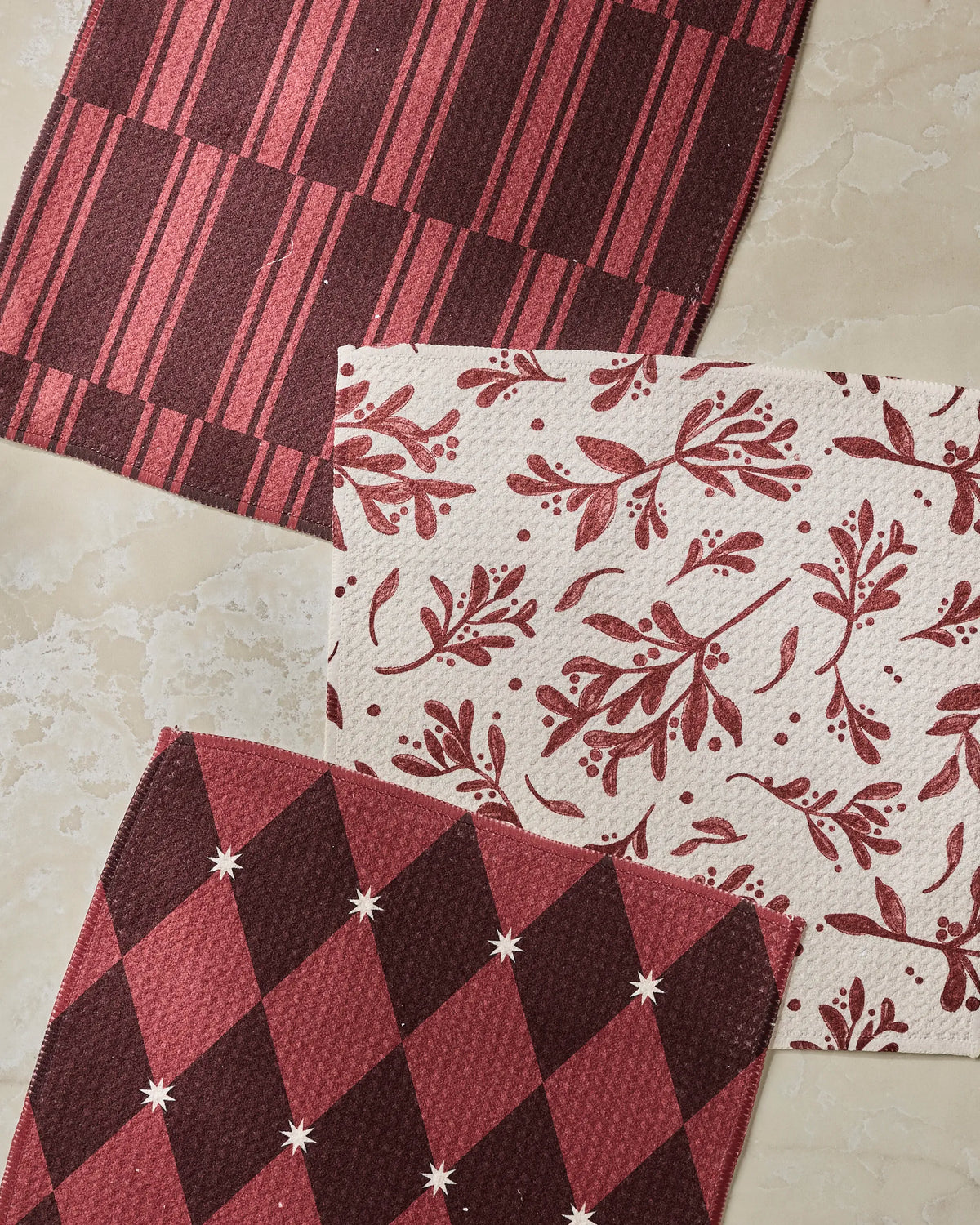 Three rectangular recycled dishcloths from Geometry’s Winter Medley collection—one with red and cream stripes, one featuring leaves and berries, and one displaying a diamond pattern—are arranged on a light marble surface. This absorbent set brings both style and sustainability to your kitchen.