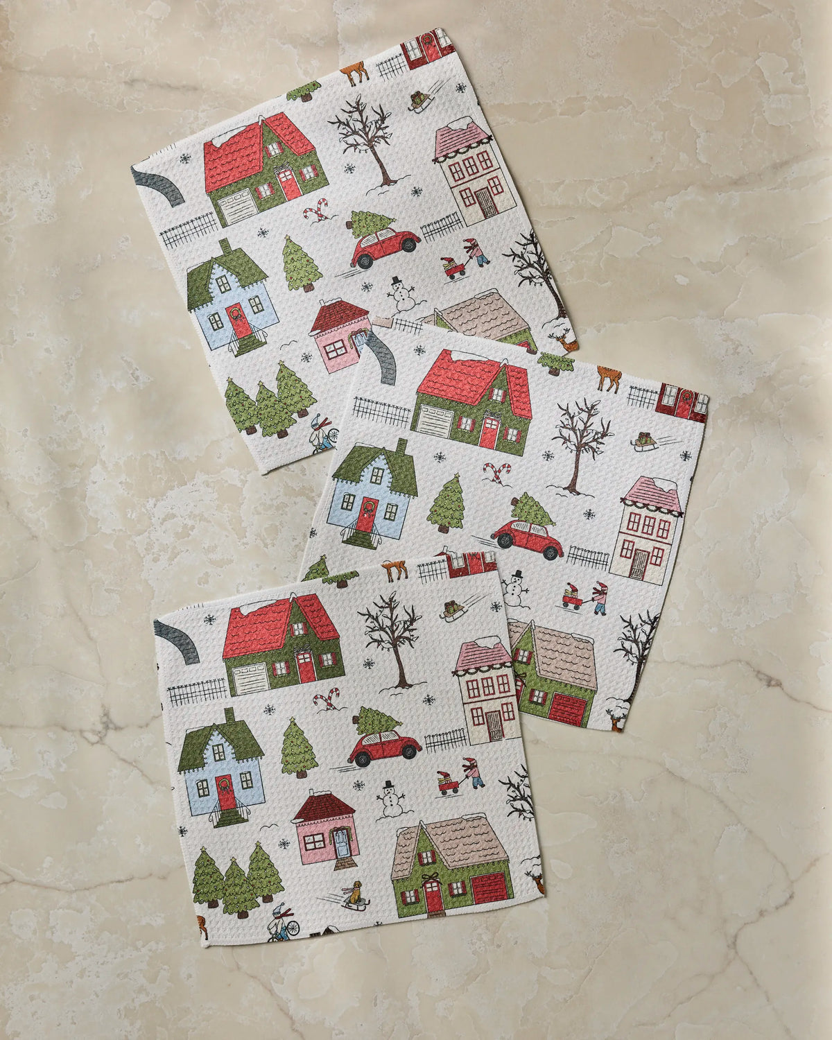 Three “Winter Village” napkins by Geometry, made from recycled materials and adorned with illustrations of houses, trees, cars, dogs, and people, are arranged on a light beige marble surface.